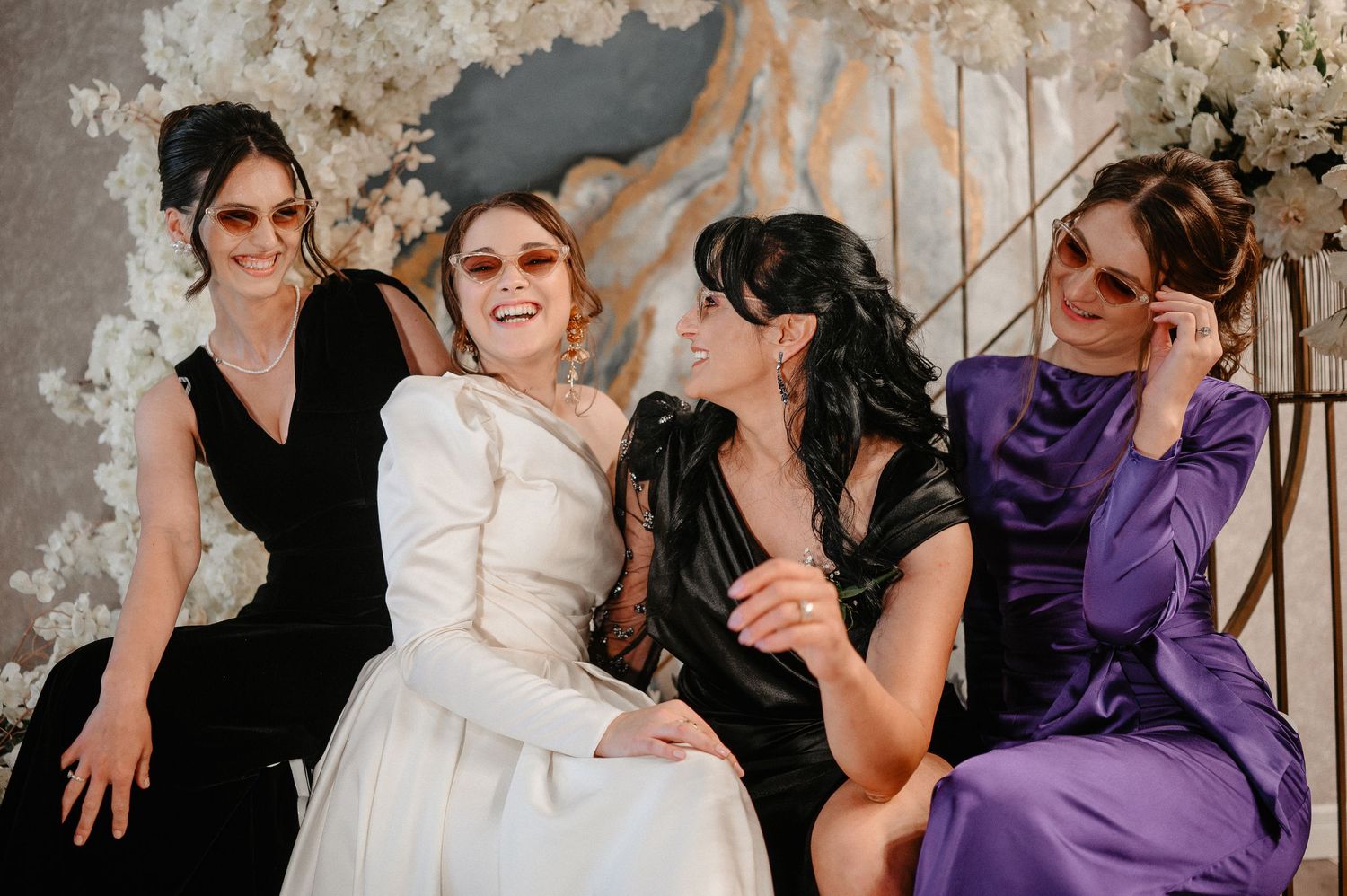 Four bridesmaids wearing sunglasses laugh together in black, white and purple dresses at wedding celebration.