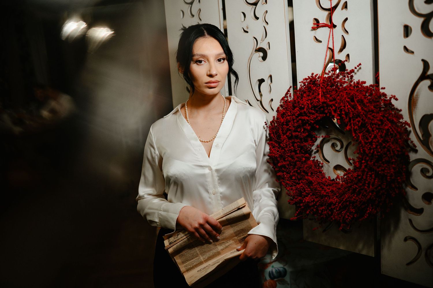 Elegant woman in white blouse holding book beside decorative red wreath on wall.