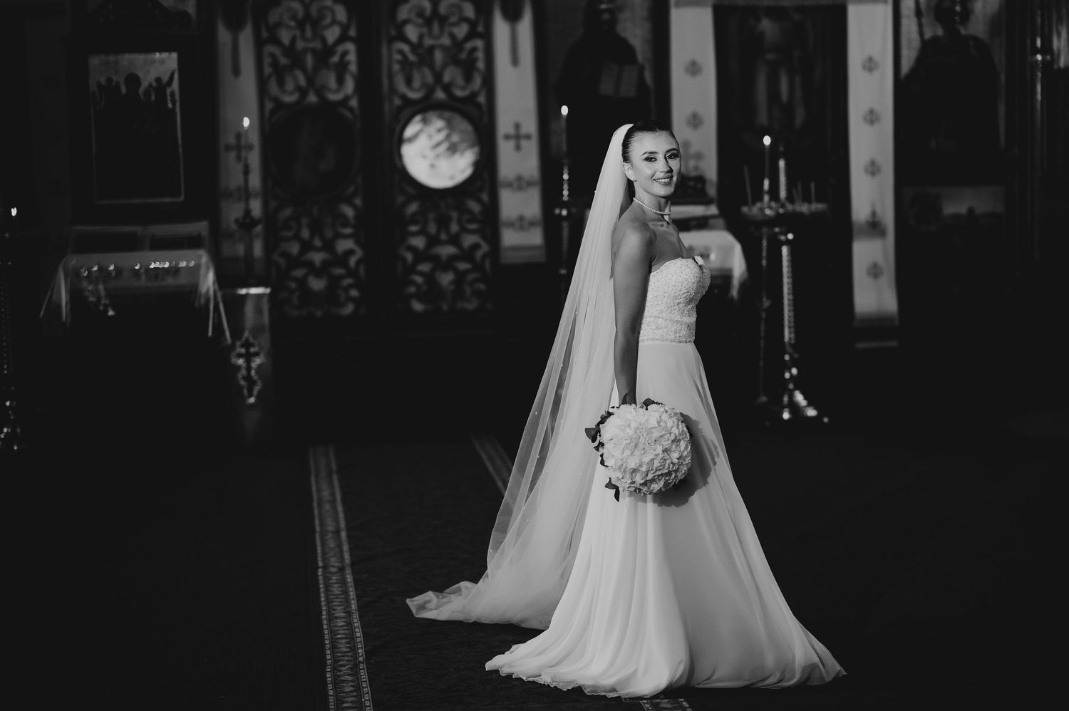 Black and white portrait of elegant bride in fitted gown and veil standing in Orthodox church.
