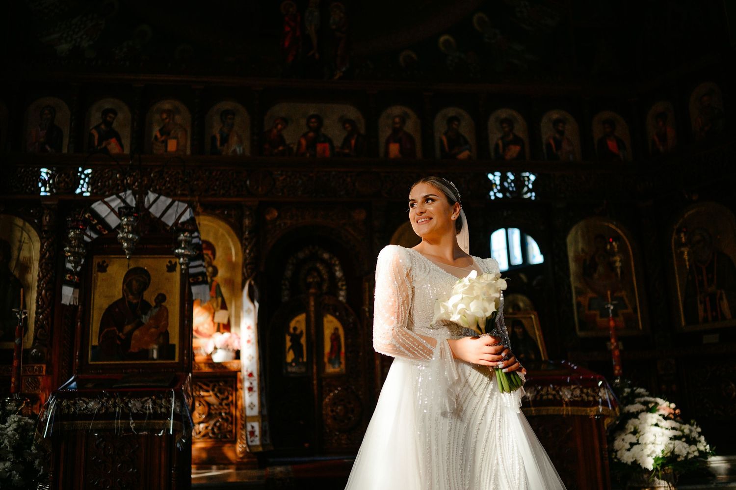 Radiant bride in beaded long-sleeve wedding gown holding white bouquet inside a beautifully decorated Orthodox church.