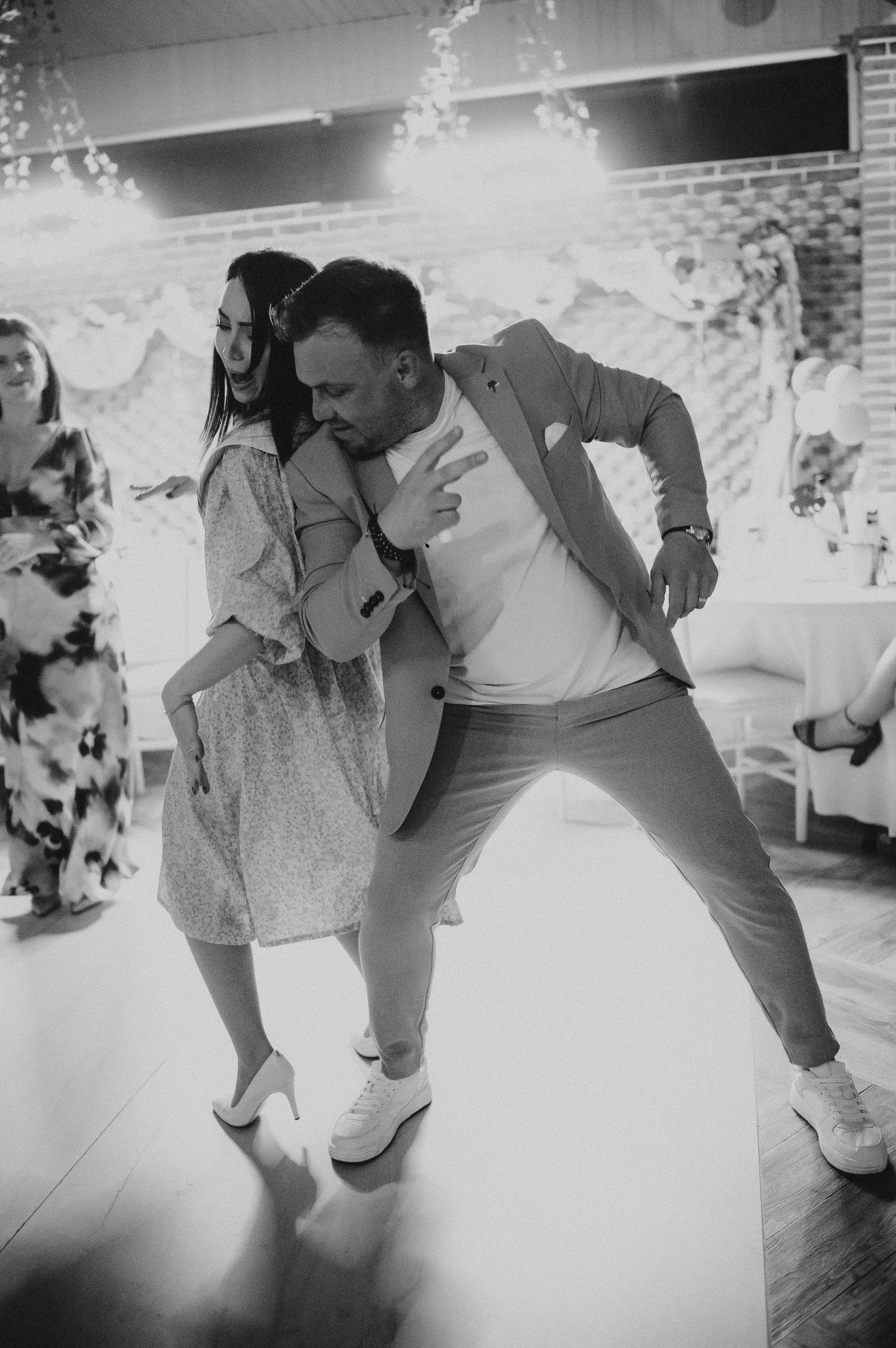Black and white photo of a man and woman dancing energetically together at an indoor celebration event.