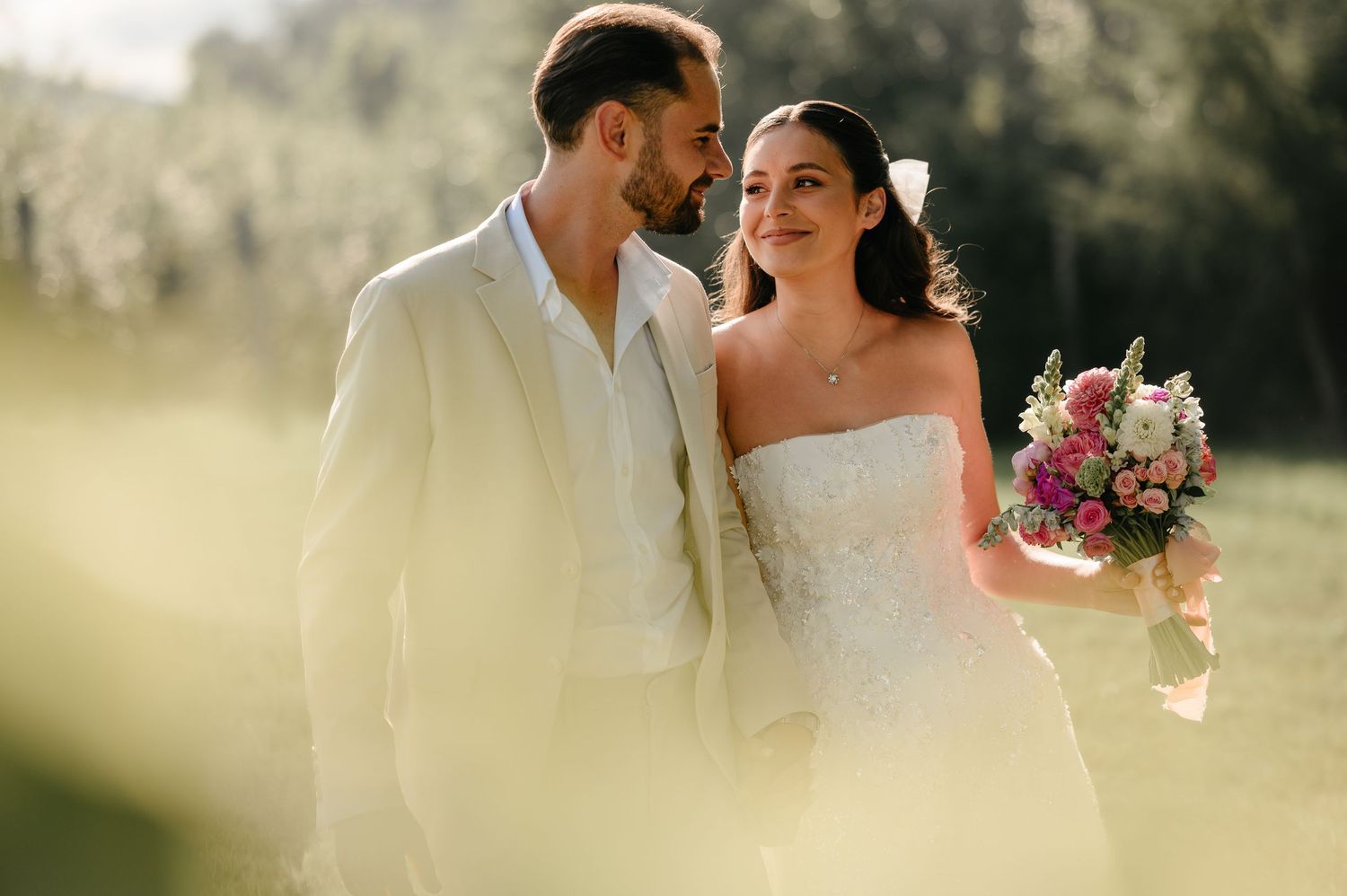 Groom in beige suit gazes at bride holding colorful bouquet in soft sunlit outdoor setting.