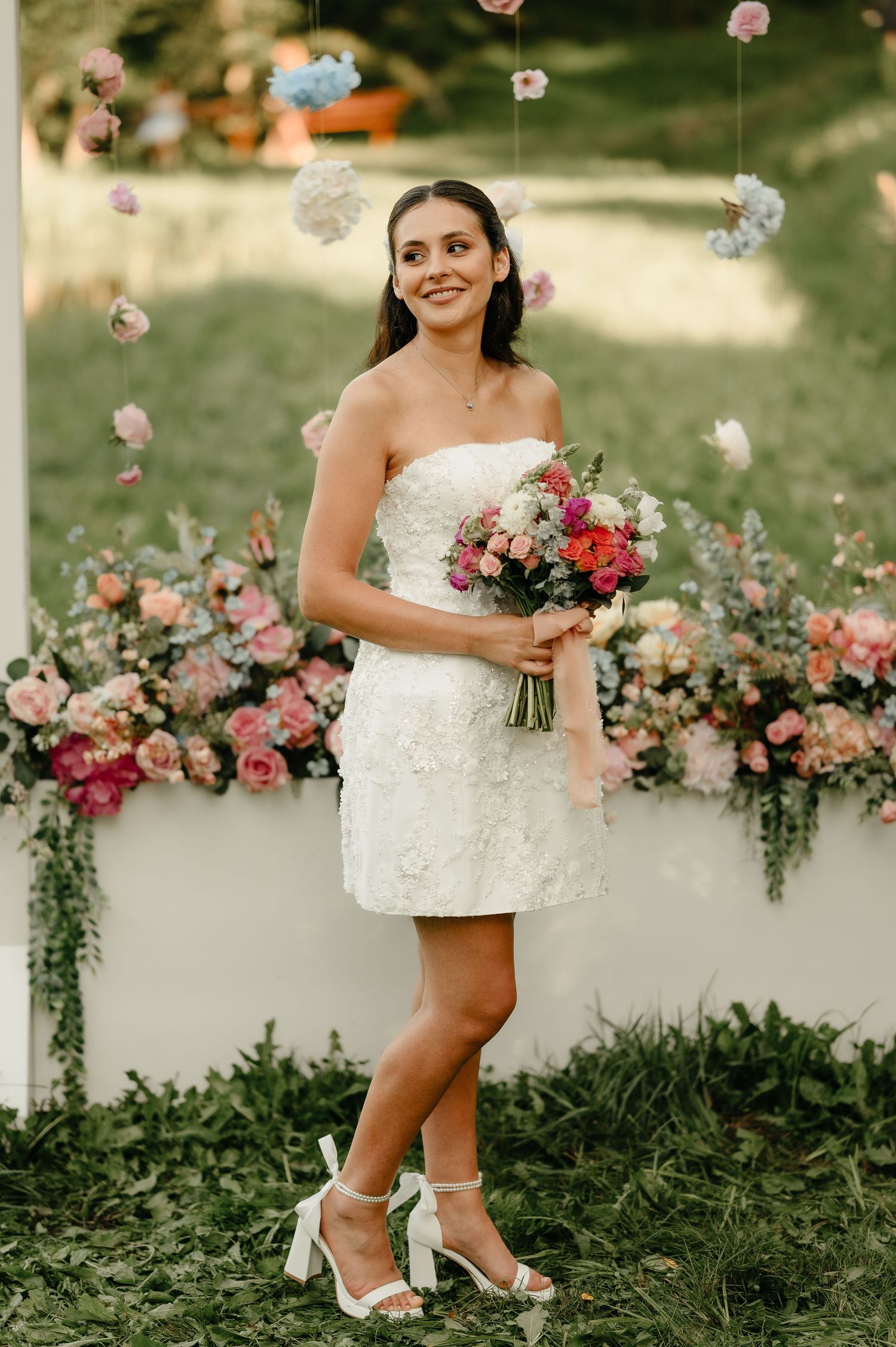 Smiling bride in short white lace dress holds colorful bouquet at floral outdoor wedding.