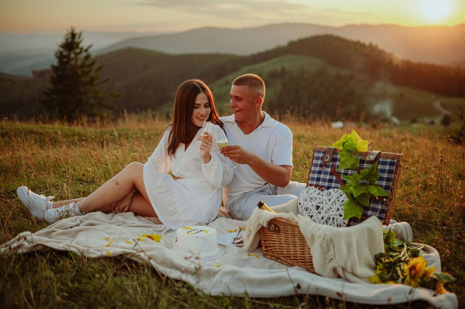 Couple enjoying a romantic picnic at sunset on a mountain hillside with wine glasses.