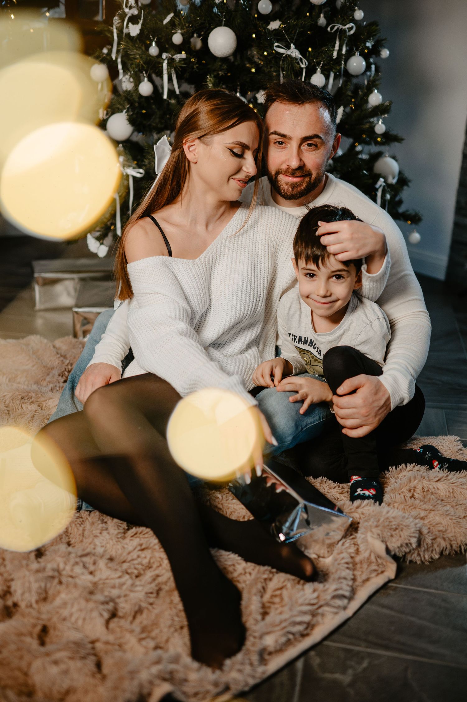 Happy family of three sitting together on fur rug by decorated Christmas tree.