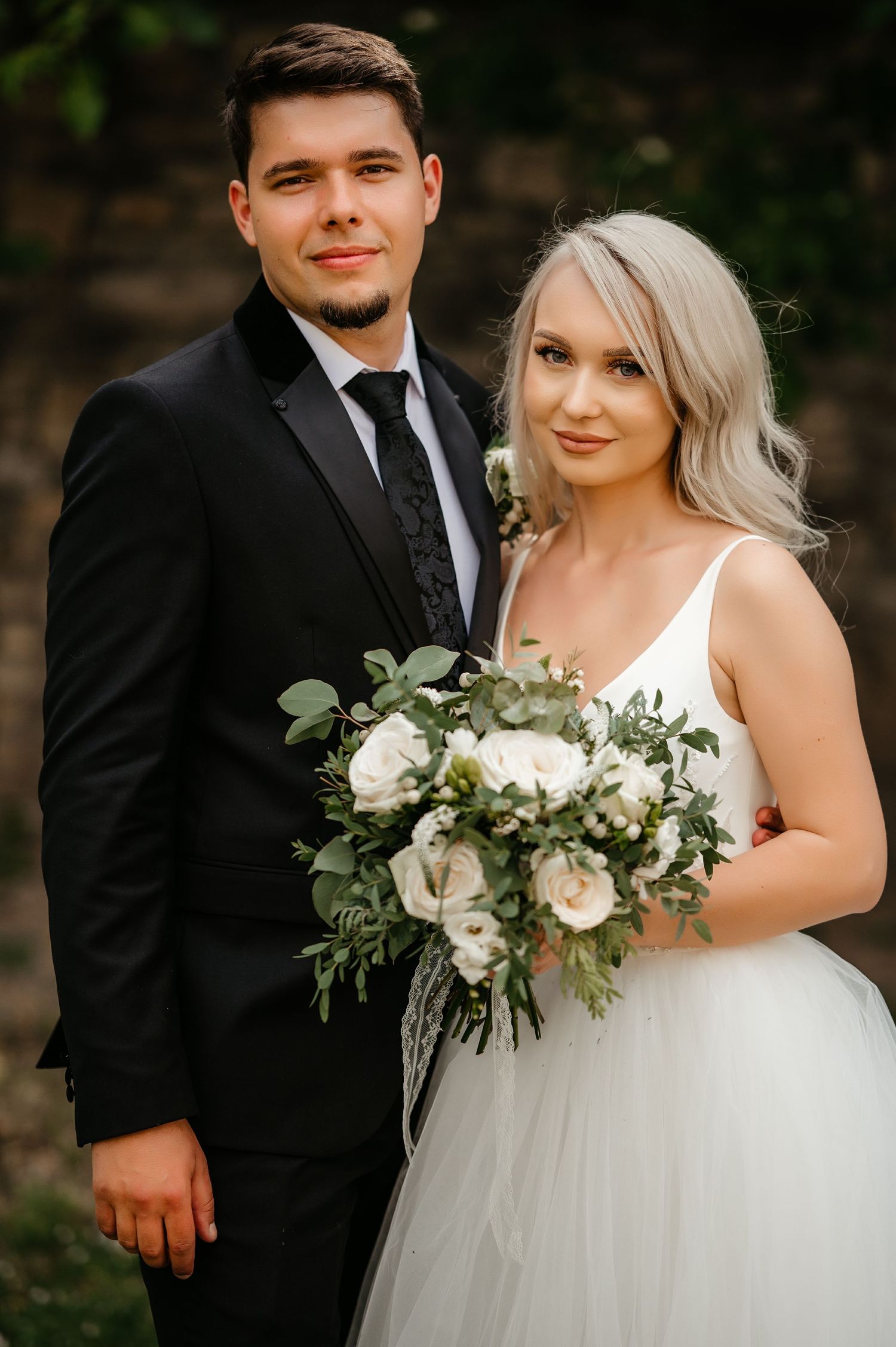 Bride holding a white and greenery bouquet poses with groom in black suit outdoors.
