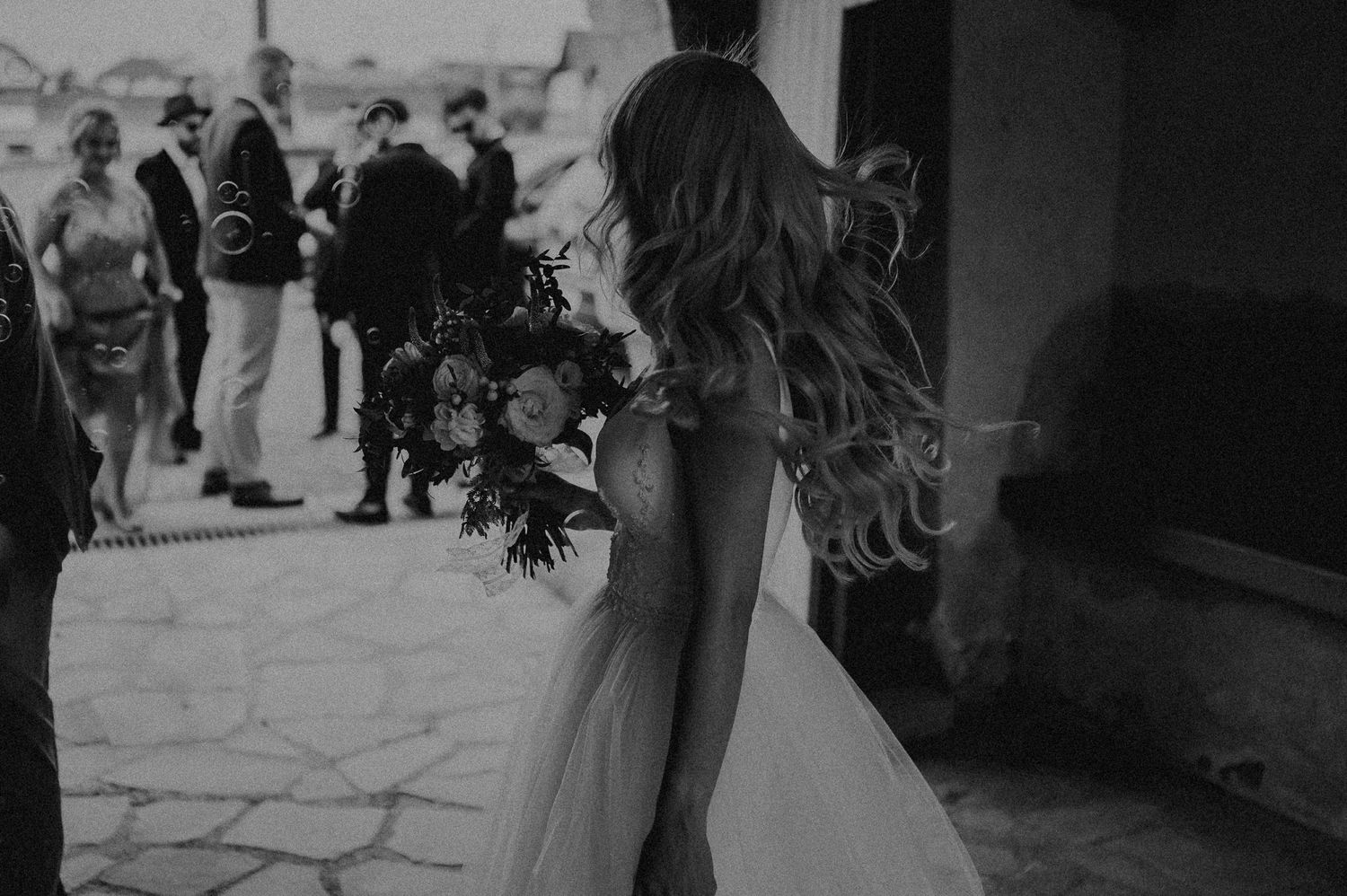 Bride in elegant backless wedding gown holding a floral bouquet in a black and white outdoor photo.