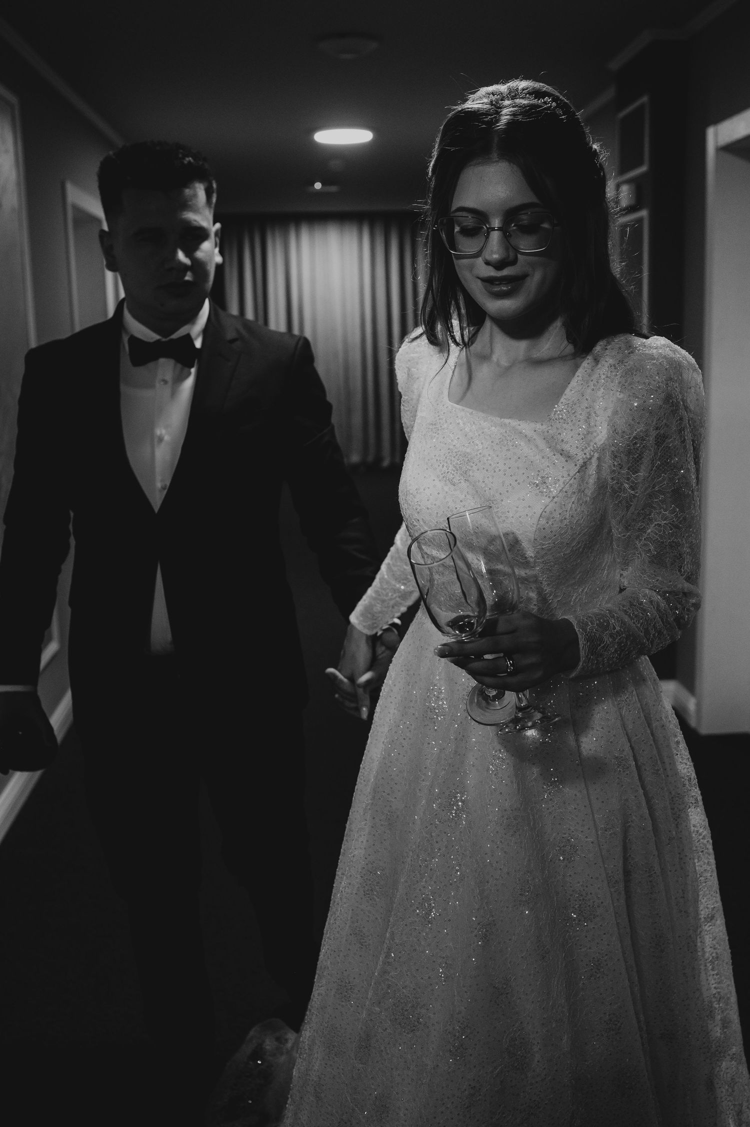 Black and white photo of bride in white gown with glasses and groom in tuxedo indoors.