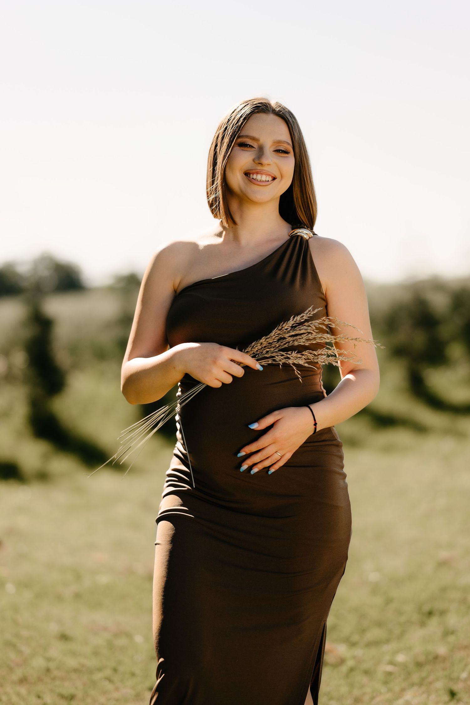 Smiling pregnant woman in elegant dark one-shoulder dress holding wheat stalks in field.