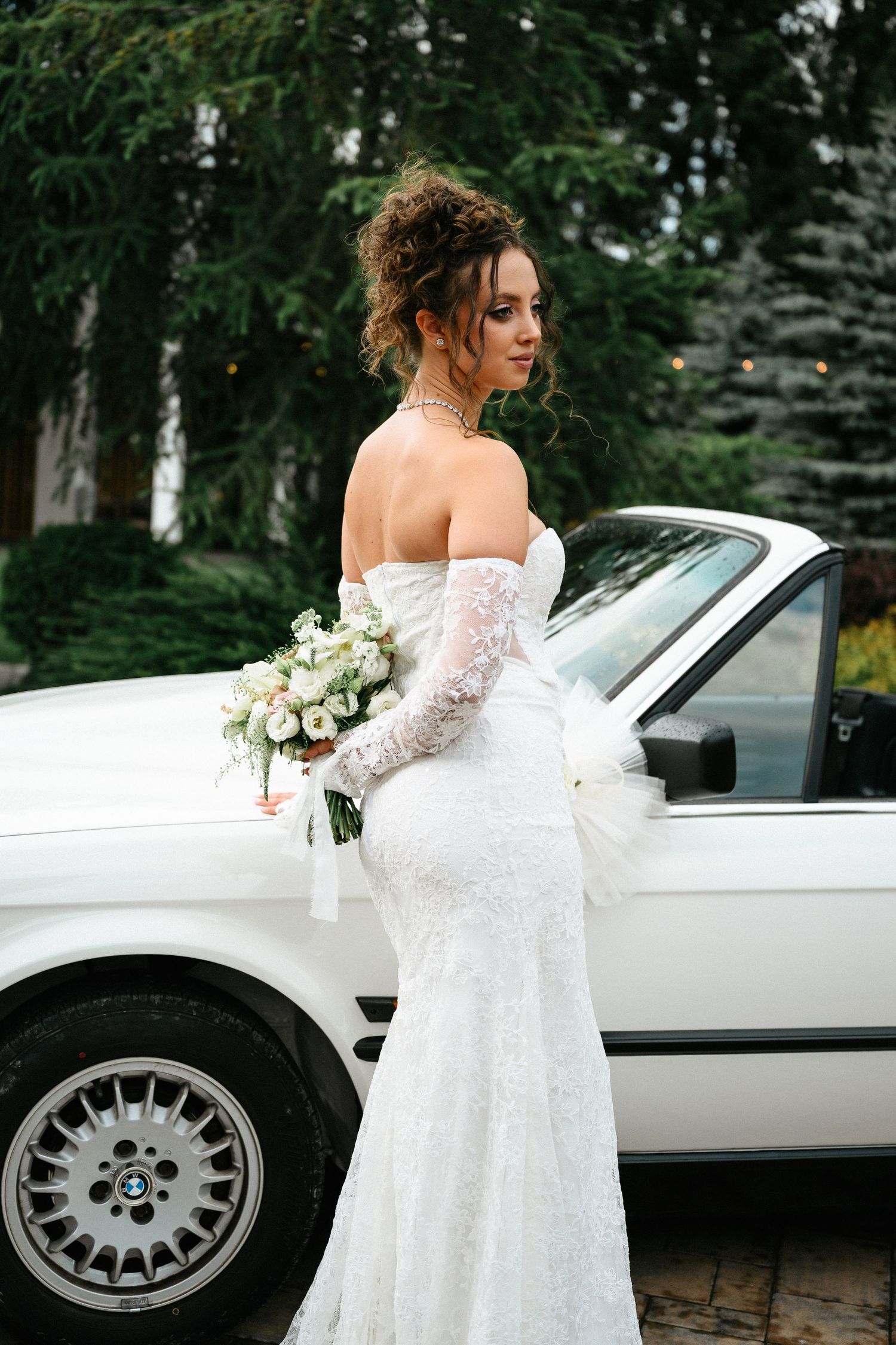 Elegant bride in white lace off-shoulder gown holding bouquet beside a classic white BMW car outdoors.