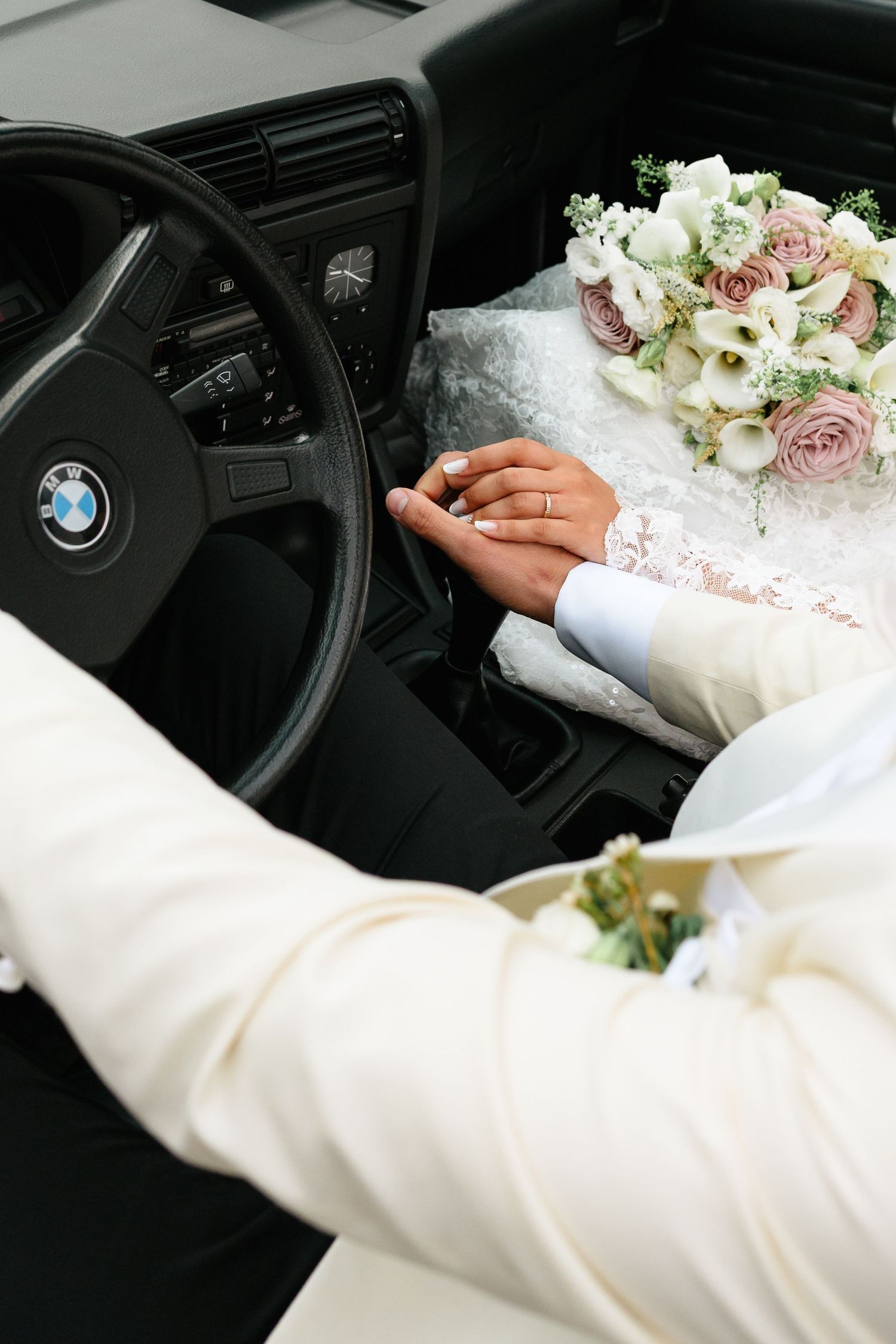 Newlyweds holding hands in a BMW car, with a bridal bouquet of pink and white roses on the dashboard.