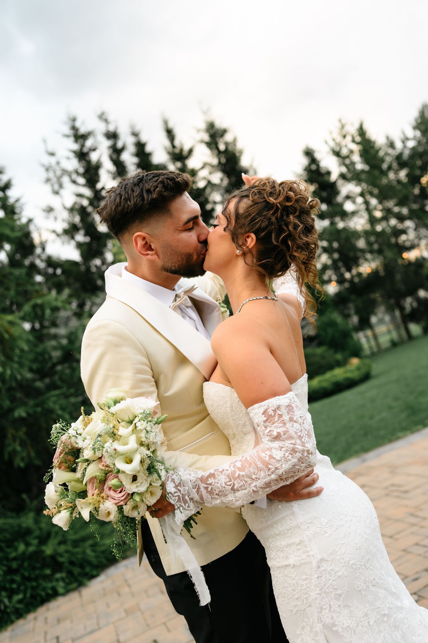 A bride and groom share a romantic kiss outdoors, she holds a bouquet in a lace gown as he wears a cream suit.