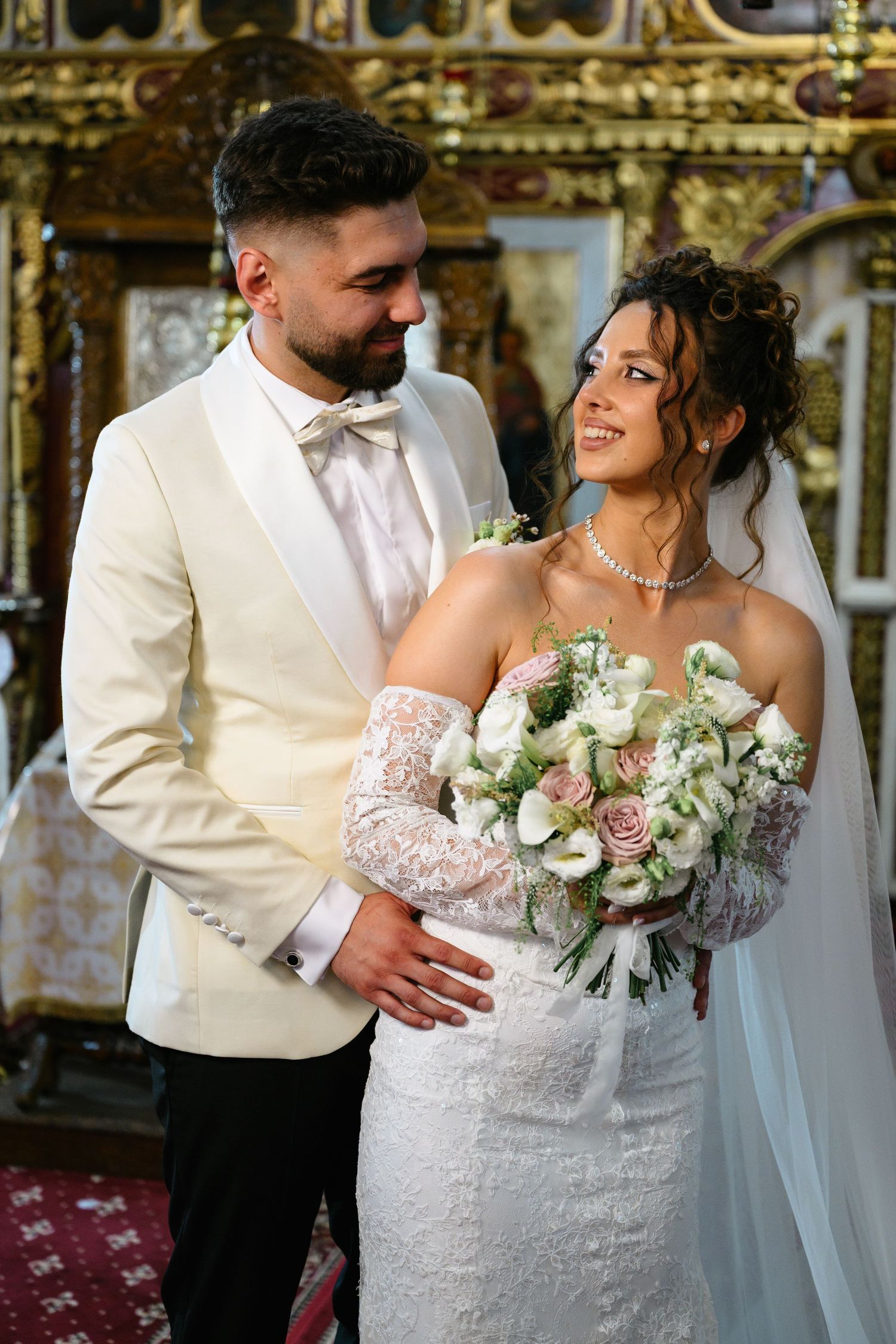 Bride and groom share a romantic moment in a church, bride holding white and pink bouquet in lace wedding dress.