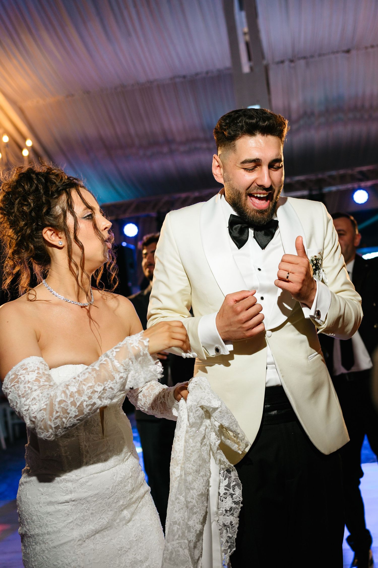 Bride and groom dancing joyfully at their wedding reception, wearing white lace gown and cream tuxedo.