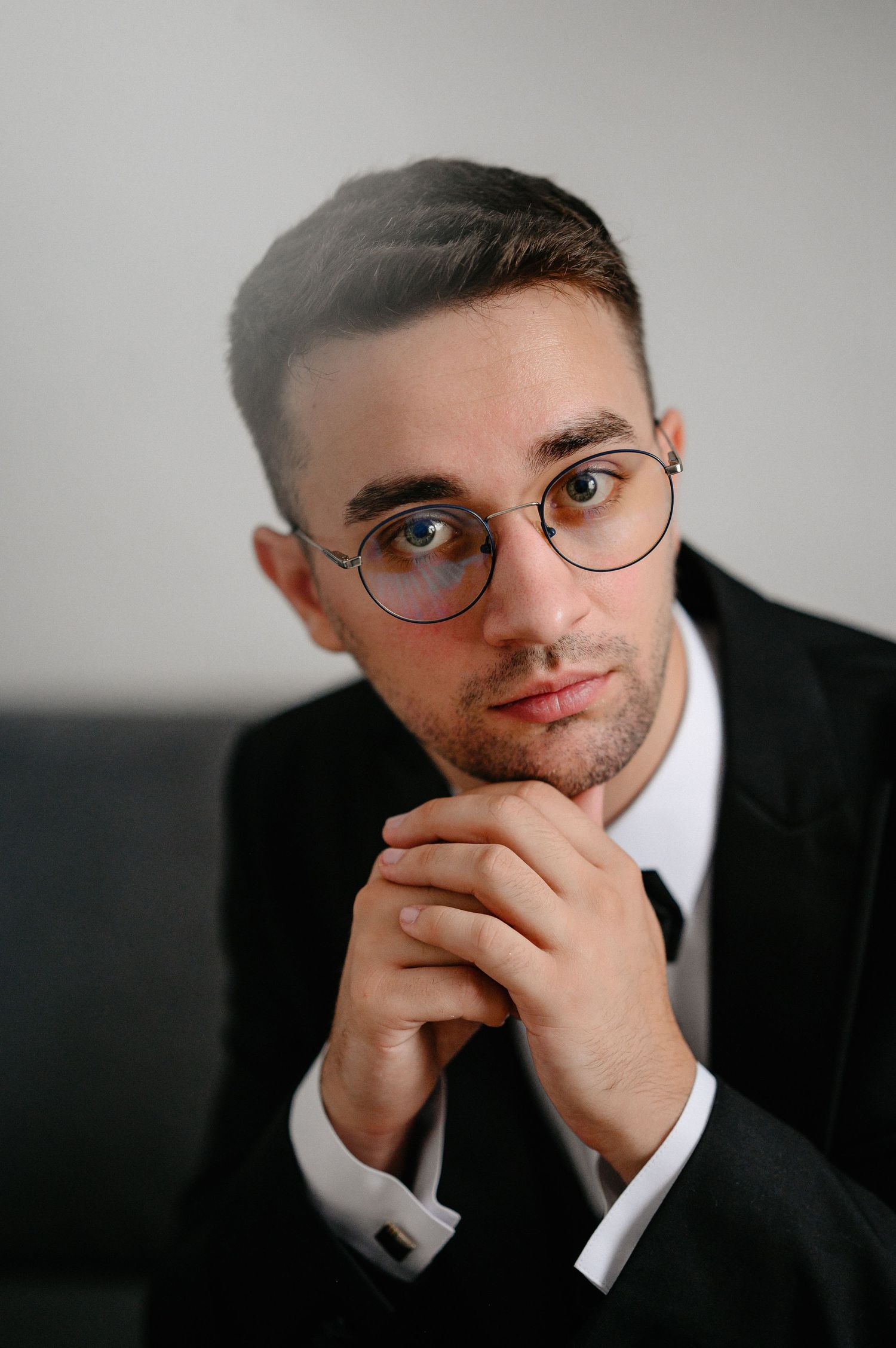 Young professional man in black suit and round glasses with hands clasped in thoughtful pose.
