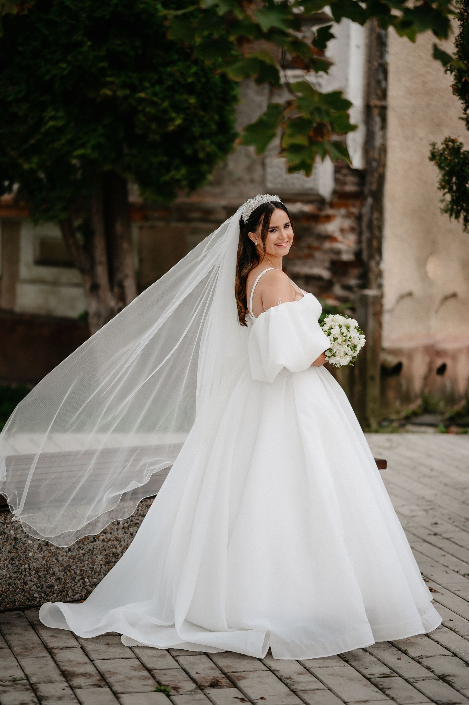 Bride in white off-shoulder ball gown with flowing veil holds bouquet outdoors near historic stone building.