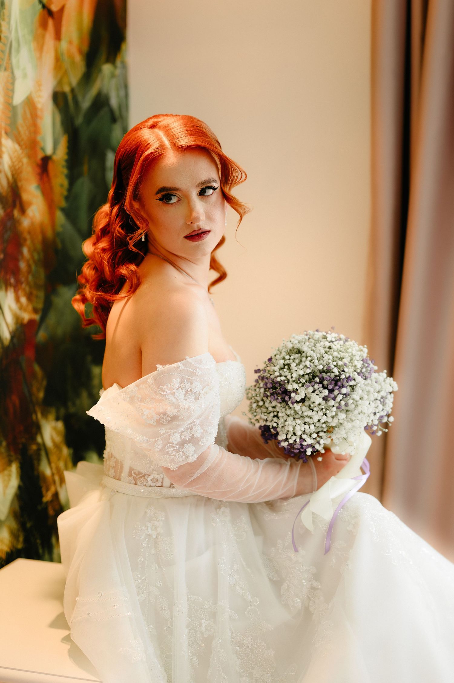 Red-haired bride in off-shoulder white lace gown holds baby's breath bouquet, posing elegantly indoors.