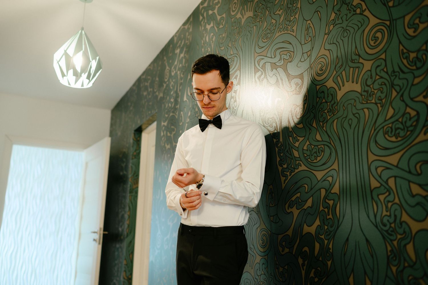 Groom in white dress shirt and bow tie adjusting cufflinks before wedding ceremony against green wallpaper.