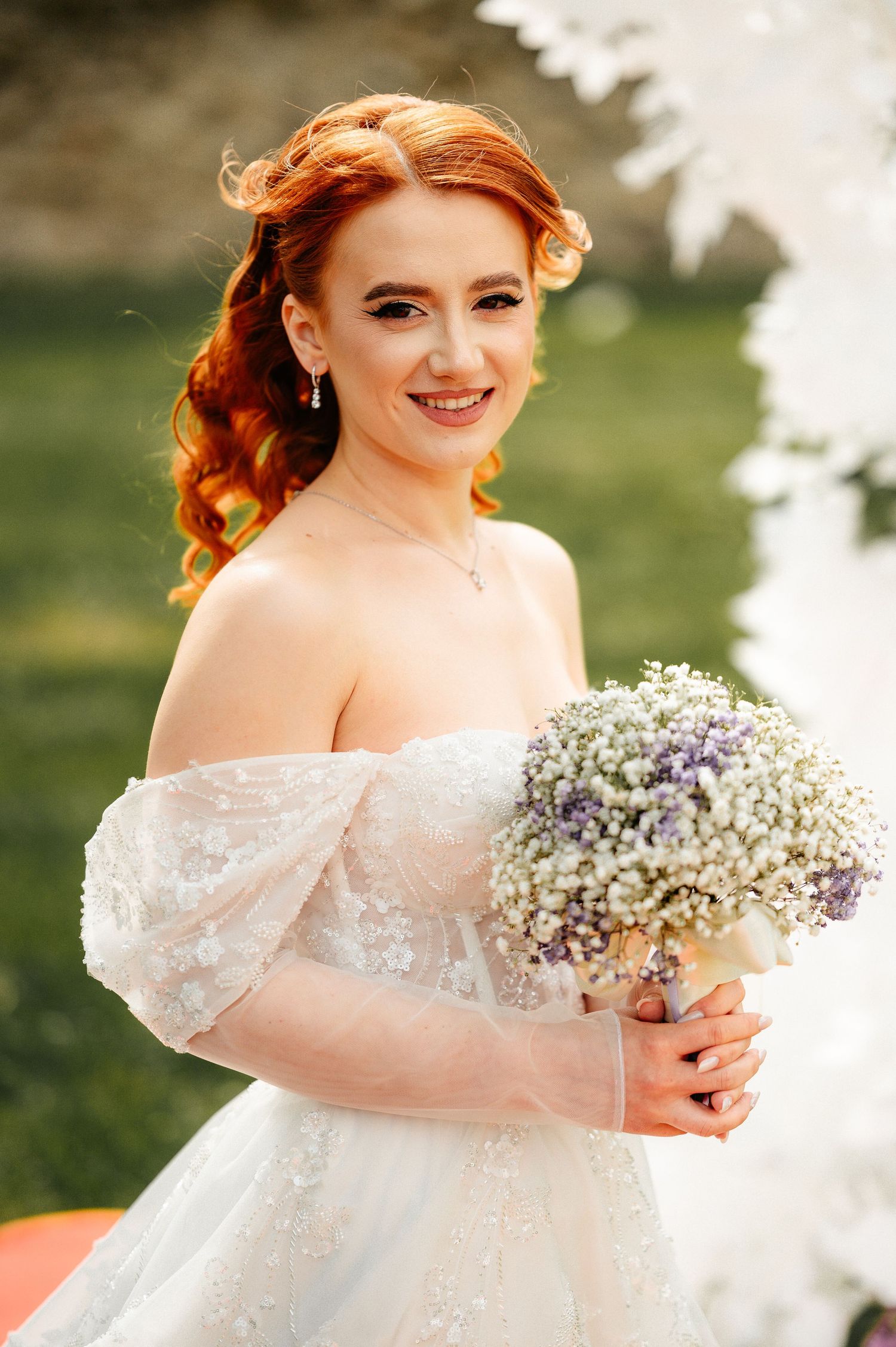 Smiling red-haired bride in off-shoulder white lace gown holding a white and purple wildflower bouquet outdoors.