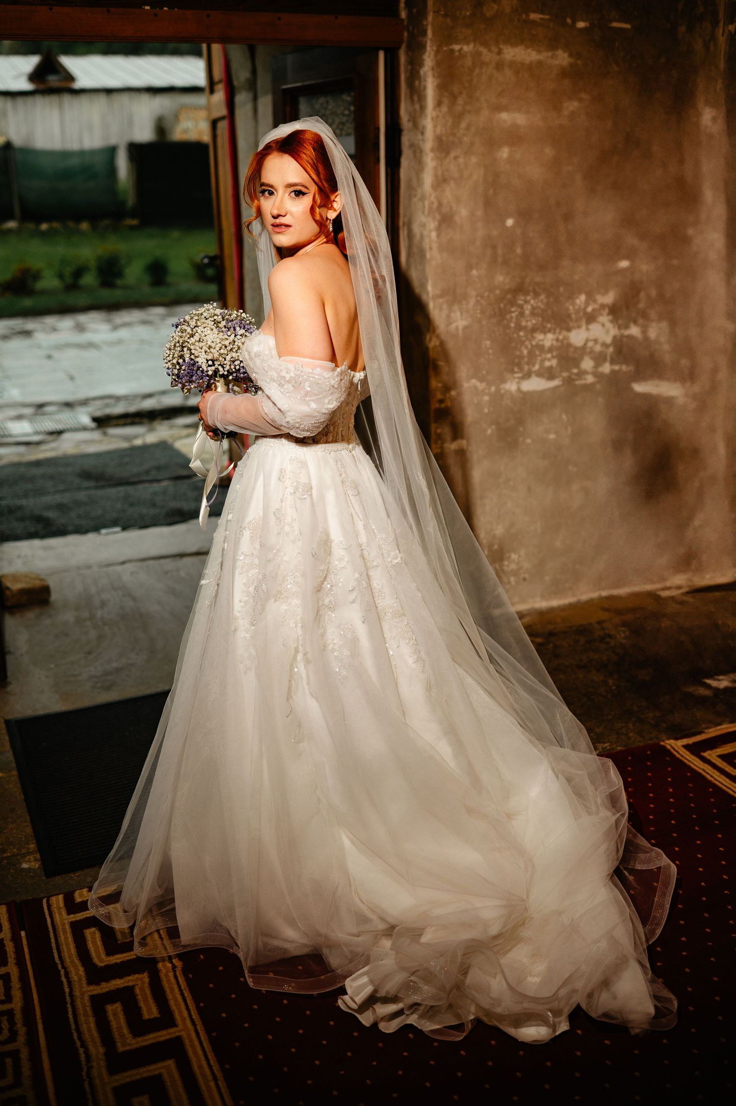 Red-haired bride in off-shoulder lace ballgown with cathedral veil holds floral bouquet in elegant venue doorway.