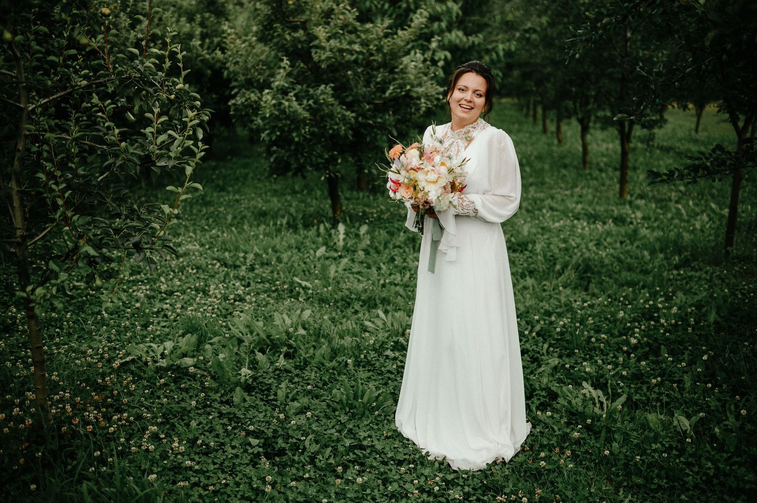 Smiling bride in white gown holding colorful bouquet standing in lush green garden surrounded by trees.