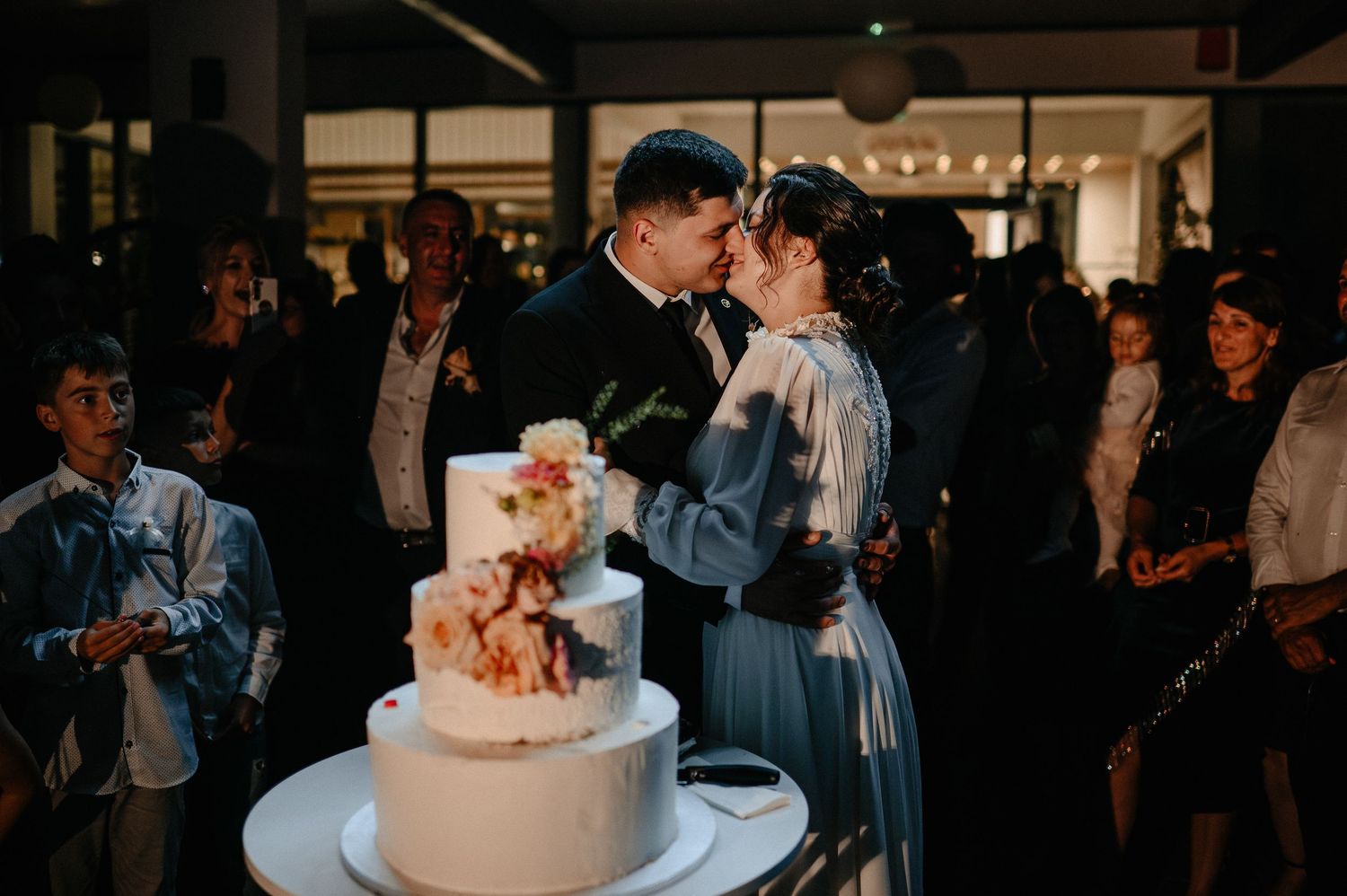 Couple kisses over elegant tiered wedding cake while guests celebrate at a stylish indoor reception.