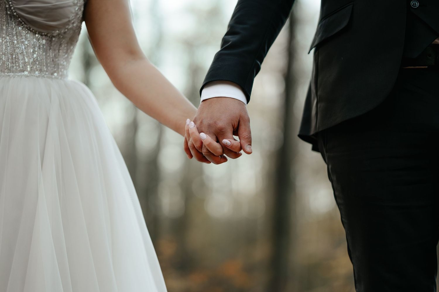 A bride and groom hold hands outdoors in a forest setting, wearing wedding attire.