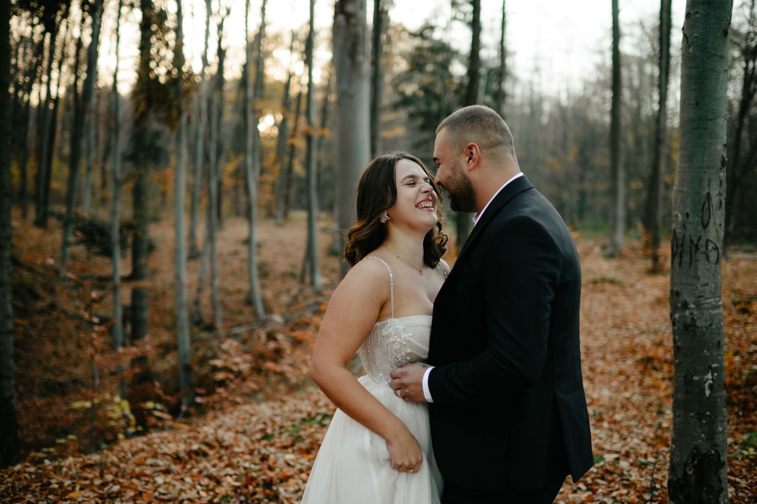Bride and groom share a romantic moment in an autumn forest, surrounded by golden fallen leaves and tall trees.