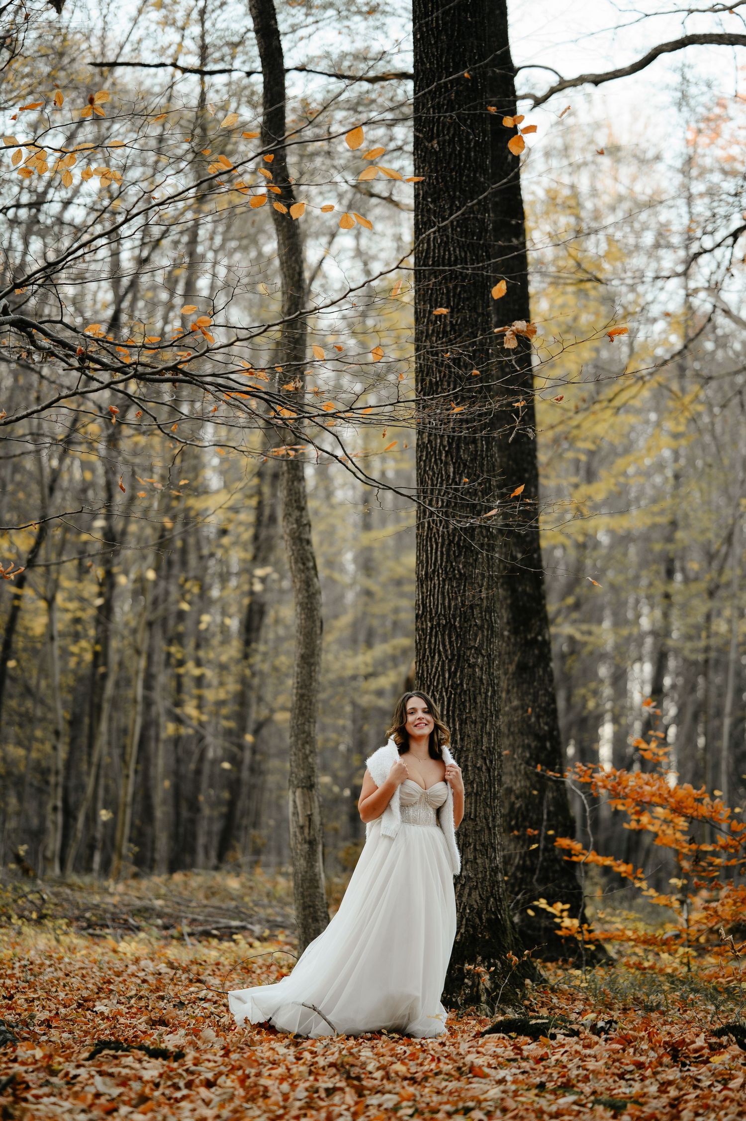 Bride in white gown leaning against large tree in autumn forest with golden fallen leaves.