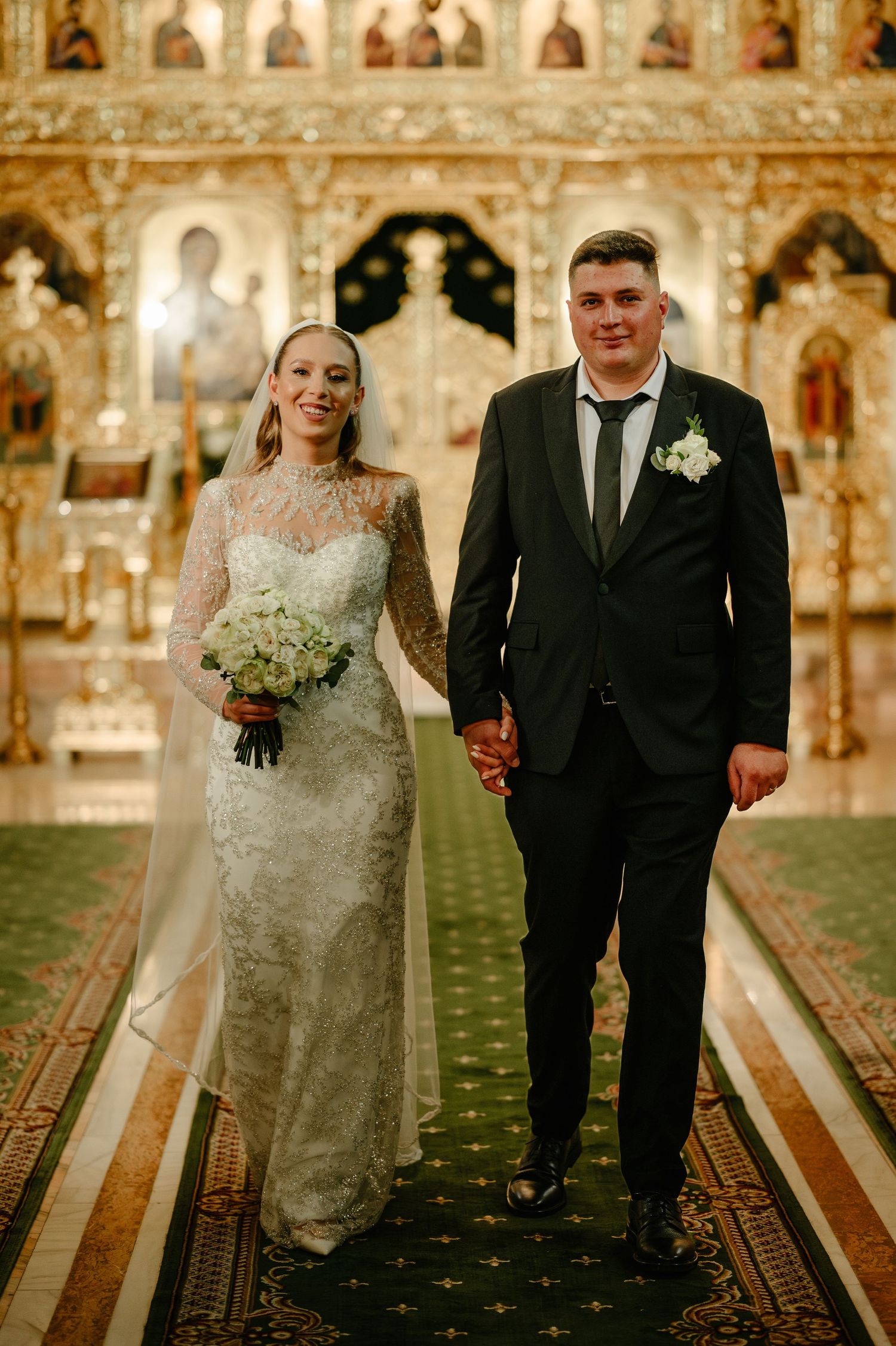 Bride and groom holding hands walking down the aisle of an ornate Orthodox church after their wedding ceremony.
