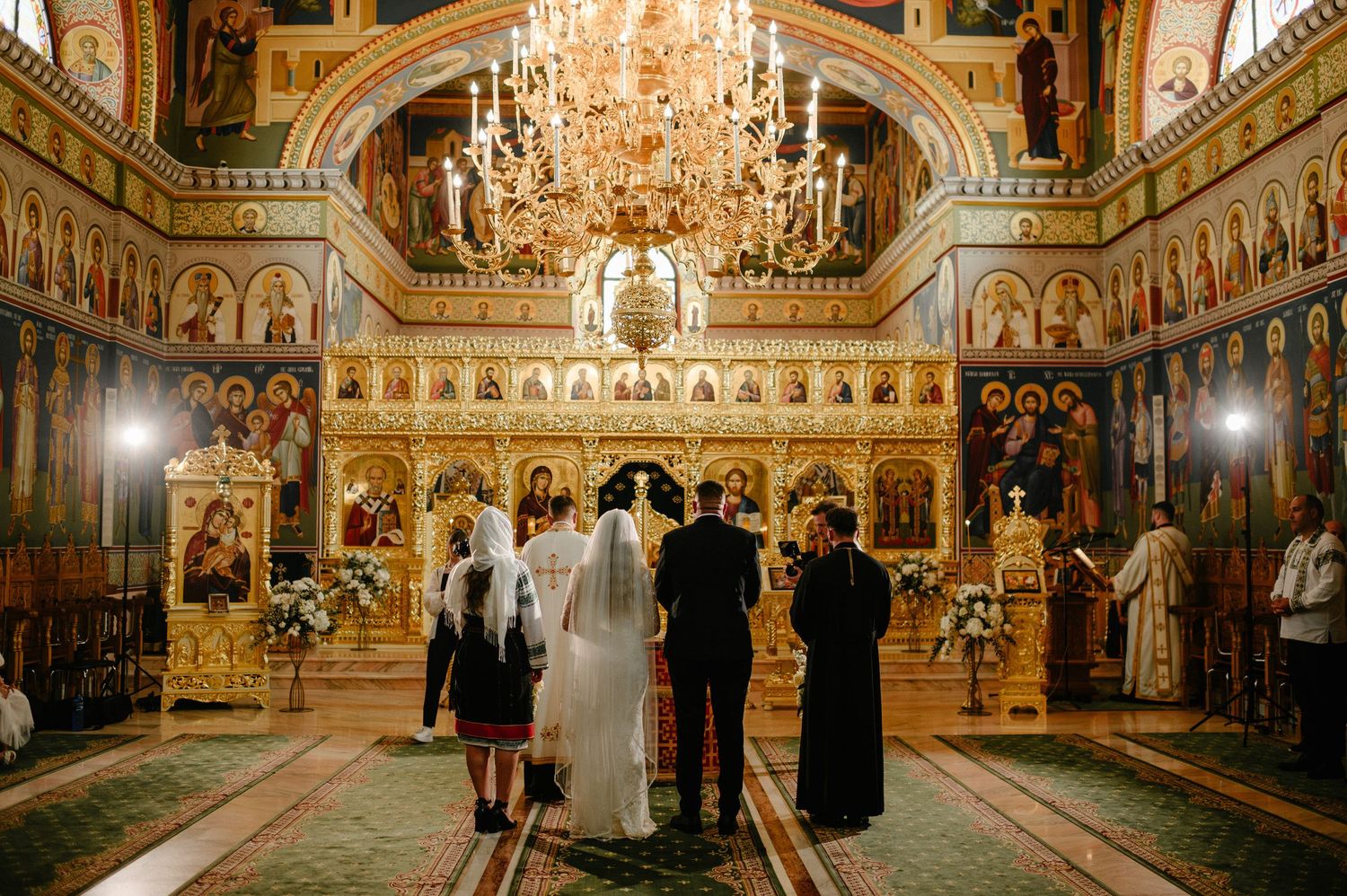 Orthodox wedding ceremony inside ornate golden church with chandelier, bride and groom standing before iconostasis.