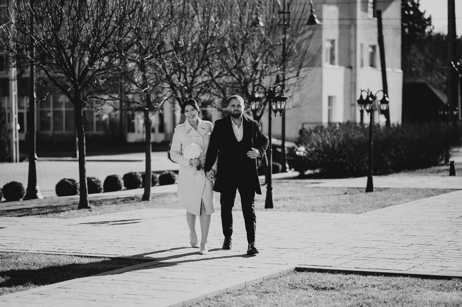 Couple walking hand in hand on a pathway in a park, dressed in elegant attire, black and white photo.