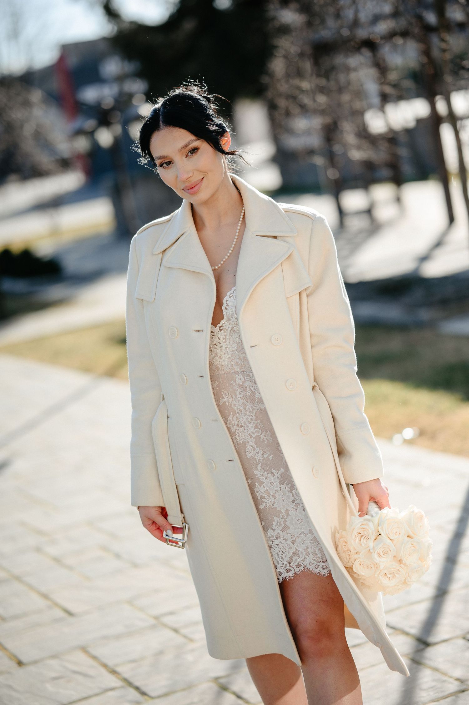 Beautiful bride smiling in ivory lace dress and long coat holding bouquet outdoors.