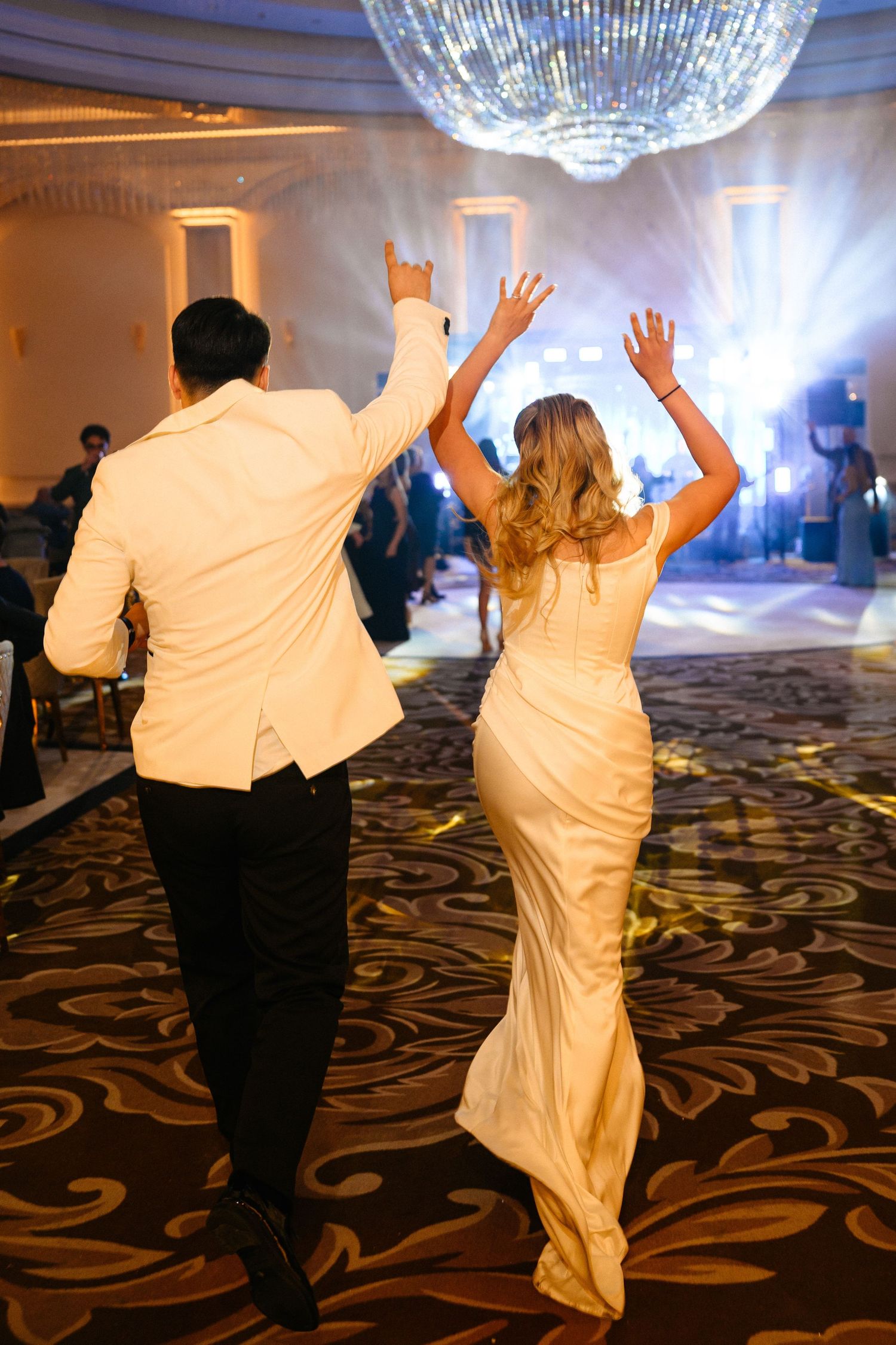 Bride and groom raising hands entering wedding reception ballroom under sparkling chandelier.