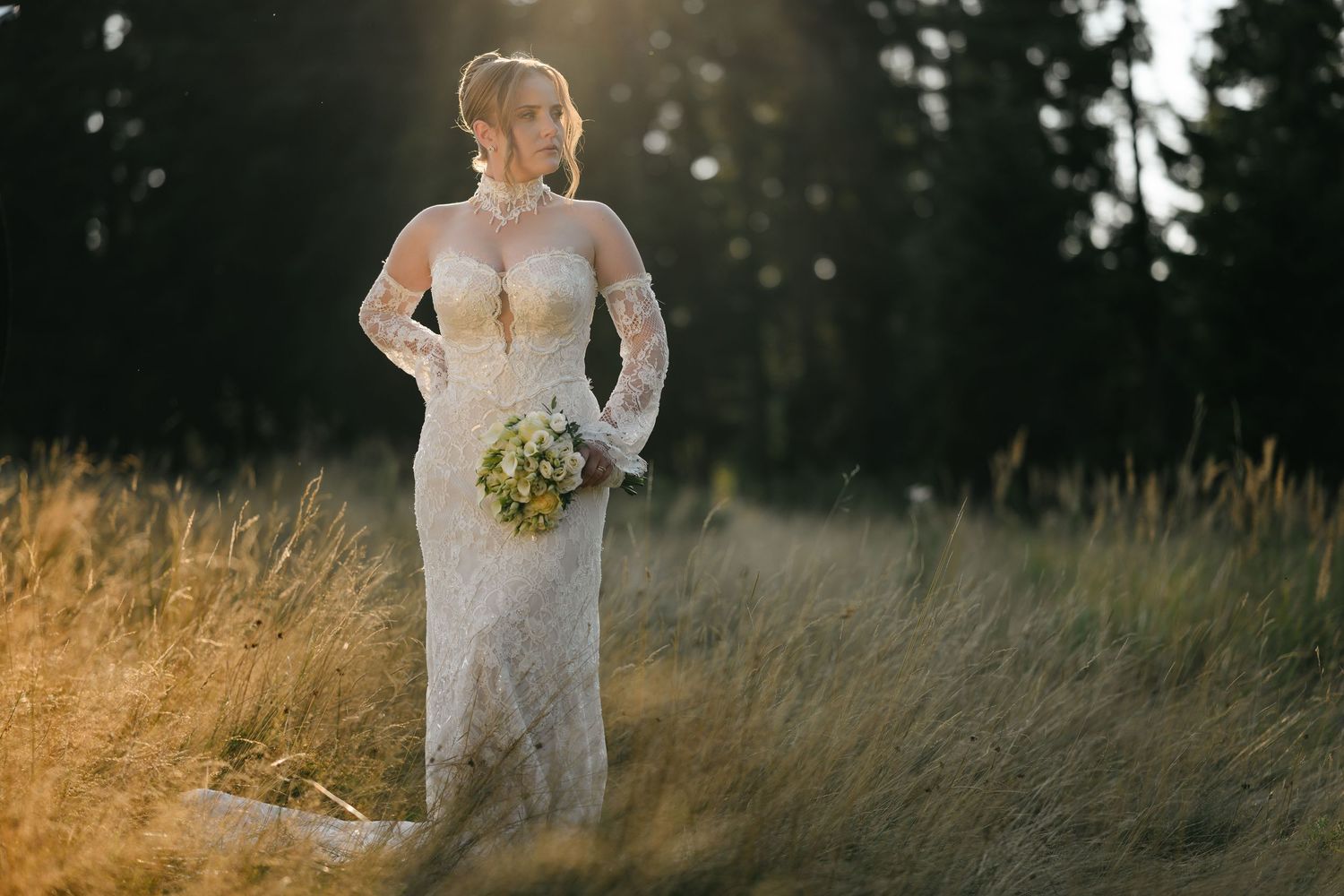 Bride in lace long-sleeve gown holding bouquet stands in golden field with sunlit forest backdrop.