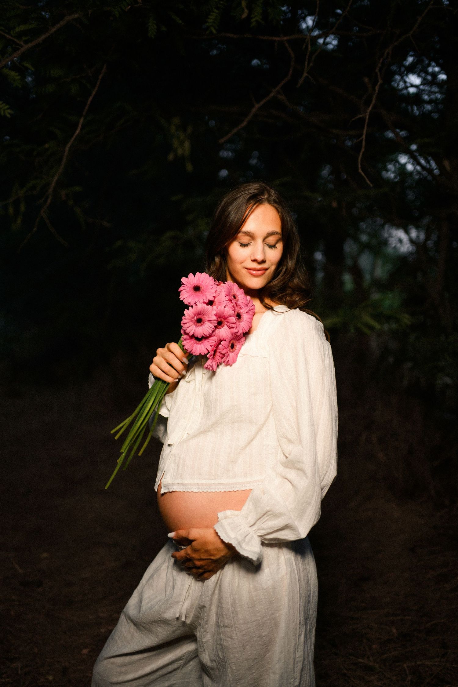 Pregnant woman in white dress holding pink gerbera daisies bouquet outdoors in soft natural light.