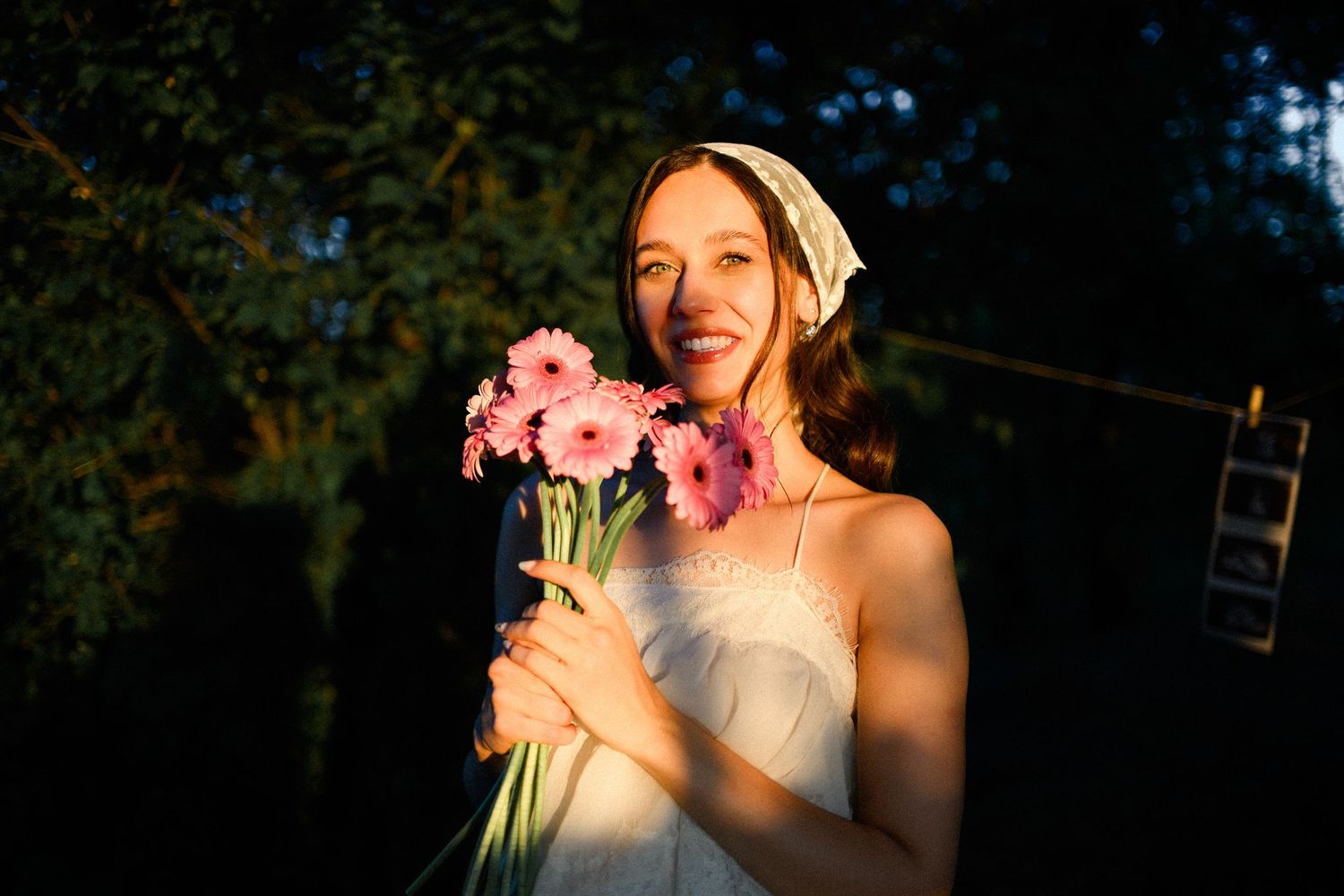 Smiling young woman in white dress holding pink gerbera daisies, bathed in warm sunset light outdoors.