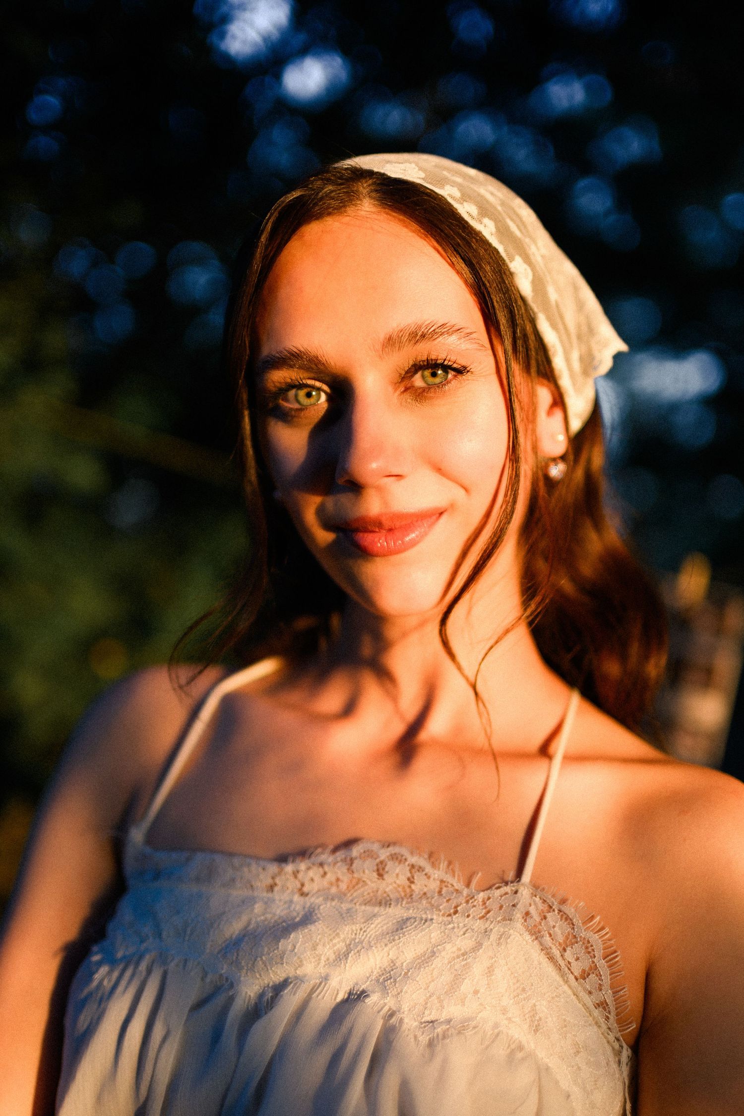 Young woman with green eyes wearing a white lace headband and dress, bathed in warm golden sunset light outdoors.