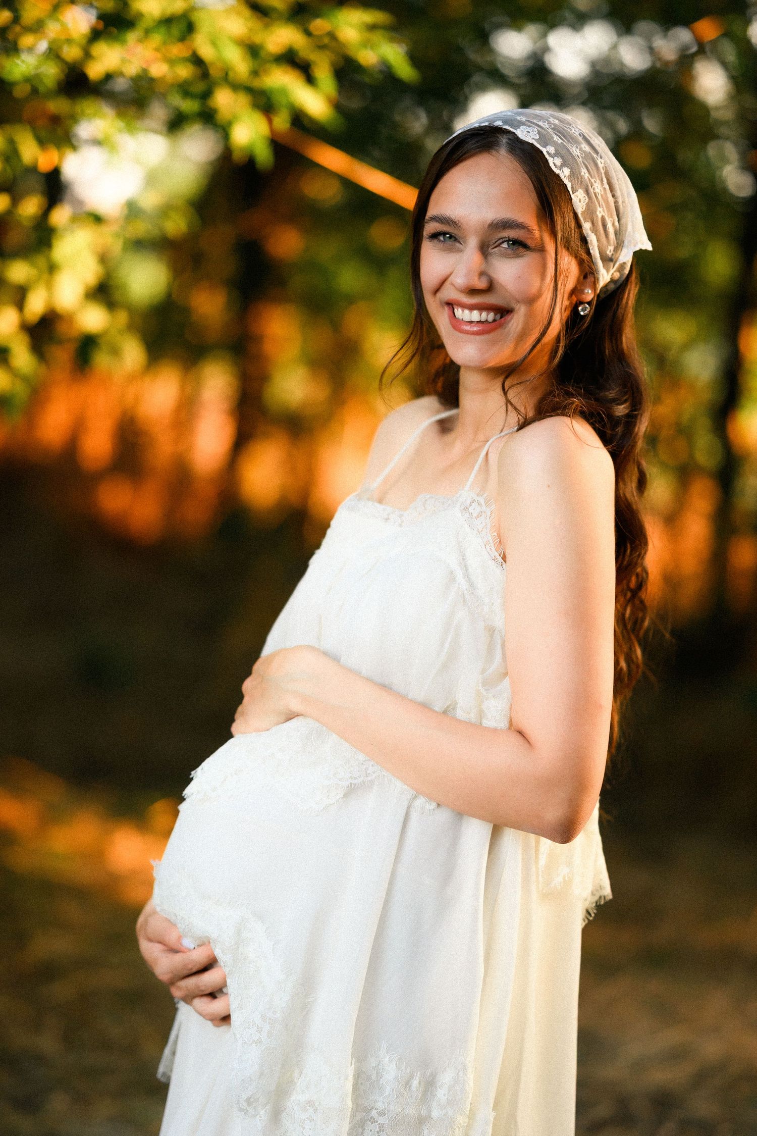 Smiling pregnant woman in white lace dress and headband cradling baby bump outdoors in golden sunlight.