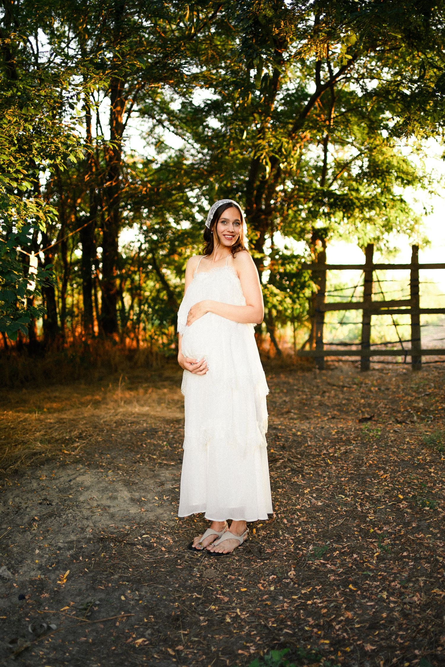 Pregnant woman in white dress standing outdoors near trees and wooden fence in golden hour light.