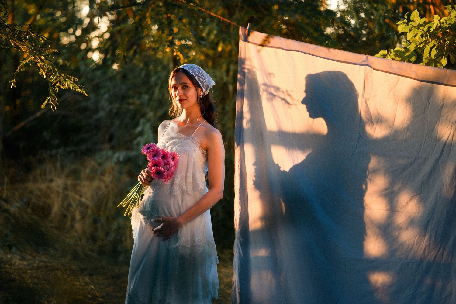 Pregnant woman in white dress holding pink flowers beside a sheer fabric panel casting her shadow in golden sunlight.