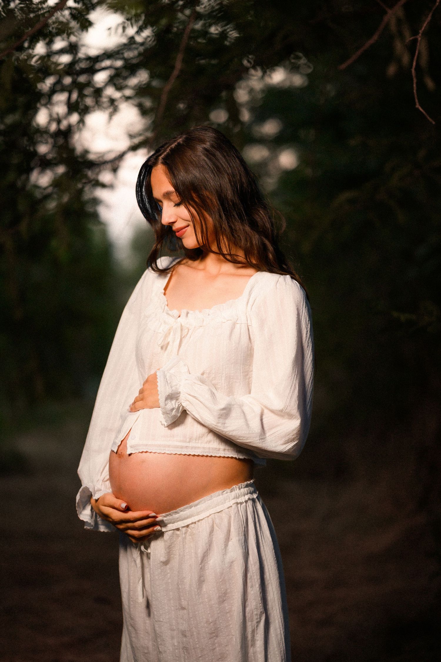 Smiling pregnant woman in white dress cradling her bare belly outdoors in soft golden sunlight among trees.