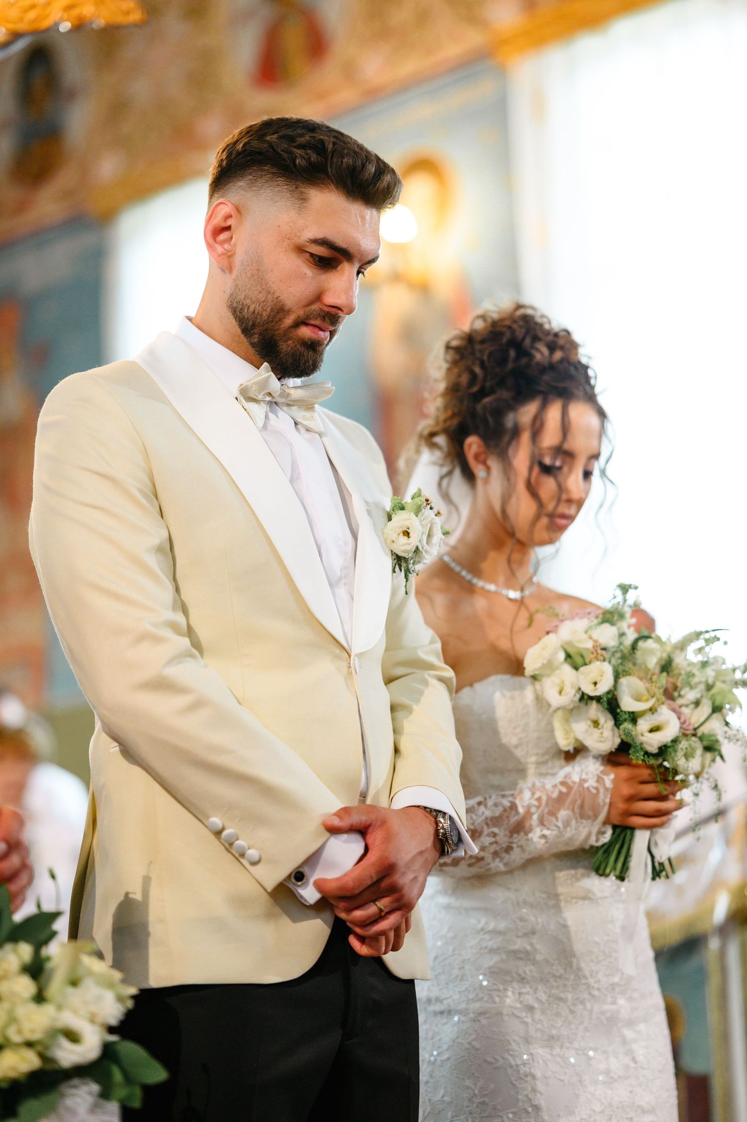 Bride and groom bow heads in prayer during elegant church wedding ceremony, groom in cream tuxedo, bride holding white bouquet.