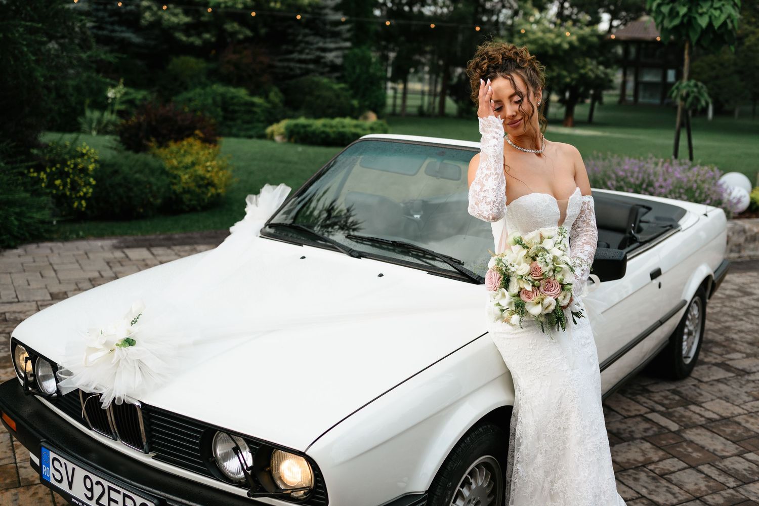 Bride in white lace gown holding bouquet beside a classic white BMW convertible decorated with ribbons outdoors.