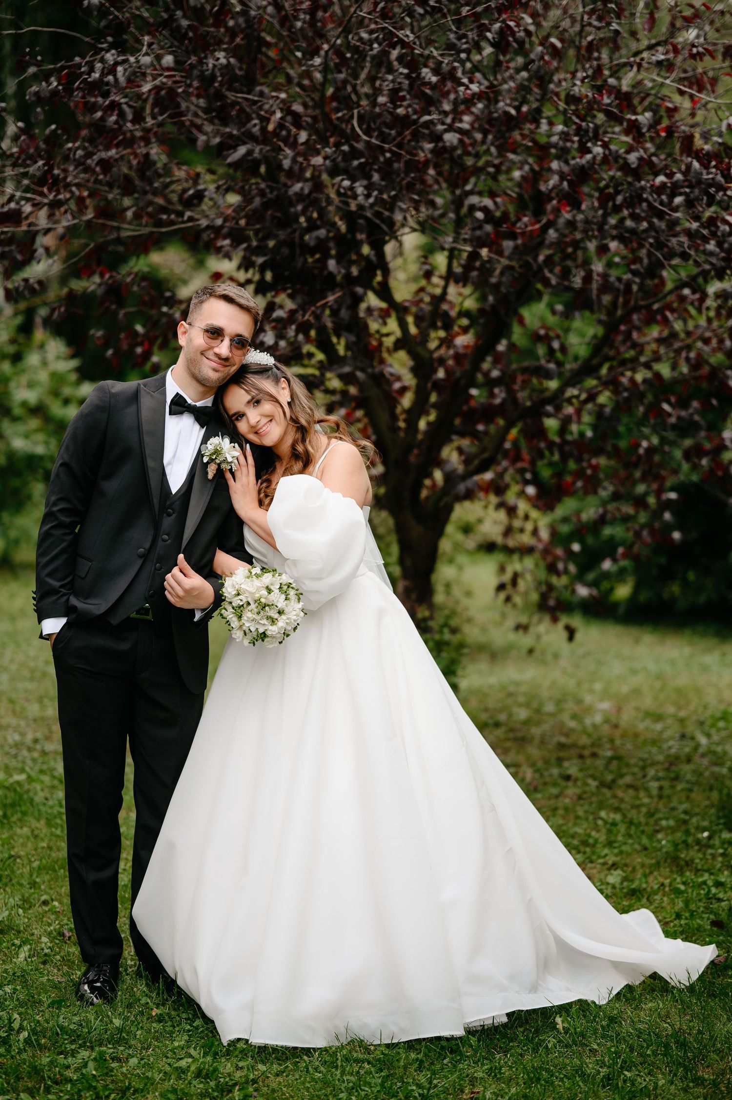 Bride in white ballgown holds bouquet leaning on groom in black tuxedo outdoors near dark foliage tree.