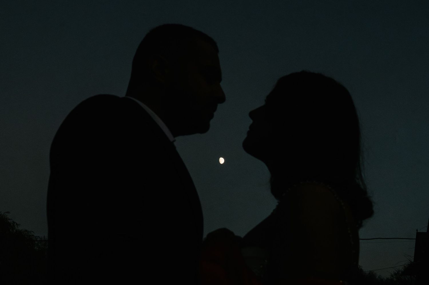 Silhouetted couple facing each other romantically at dusk with a glowing moon between them against a dark blue sky.