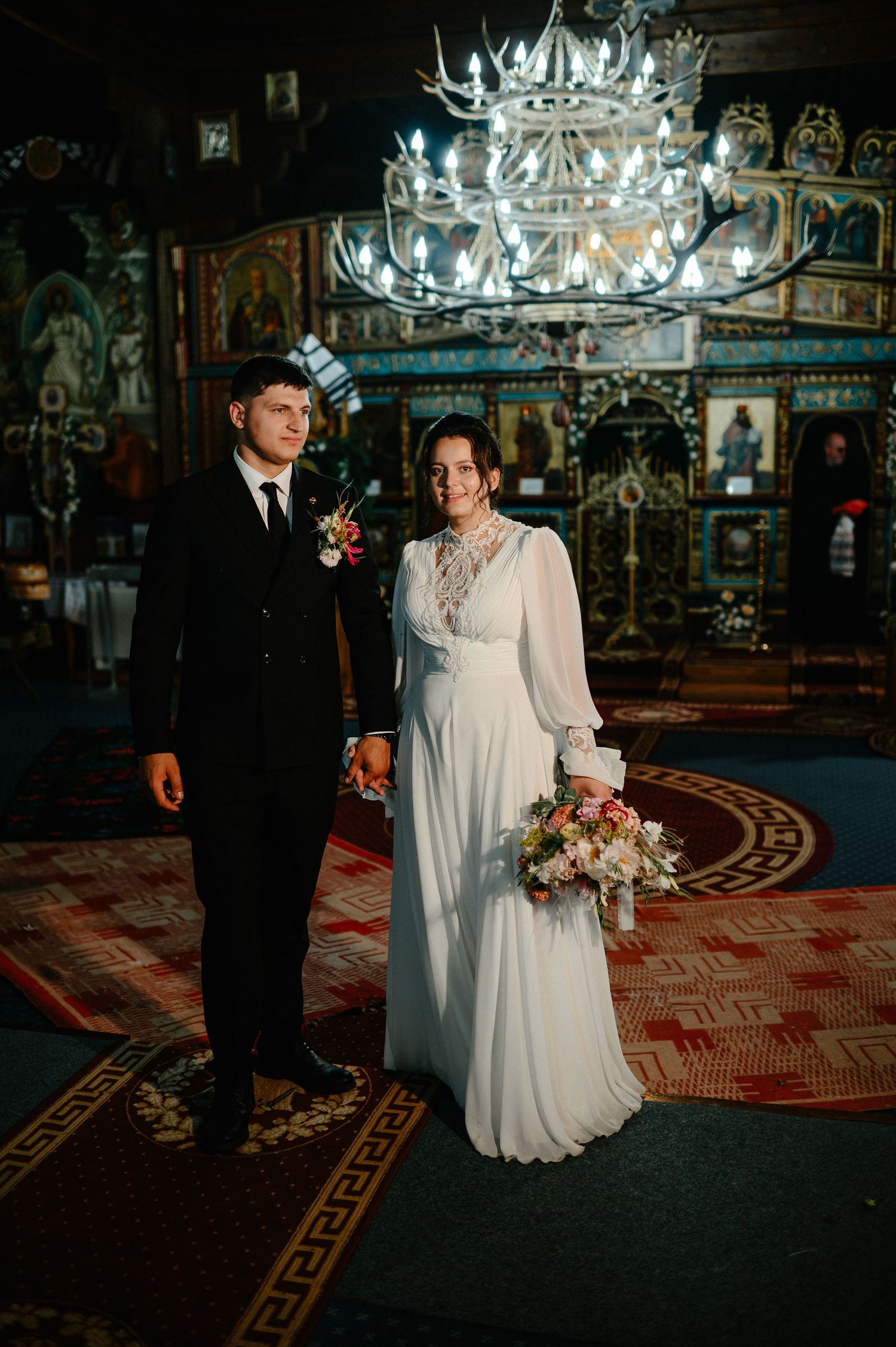 Bride and groom stand hand in hand inside ornate Orthodox church with antler chandelier overhead.