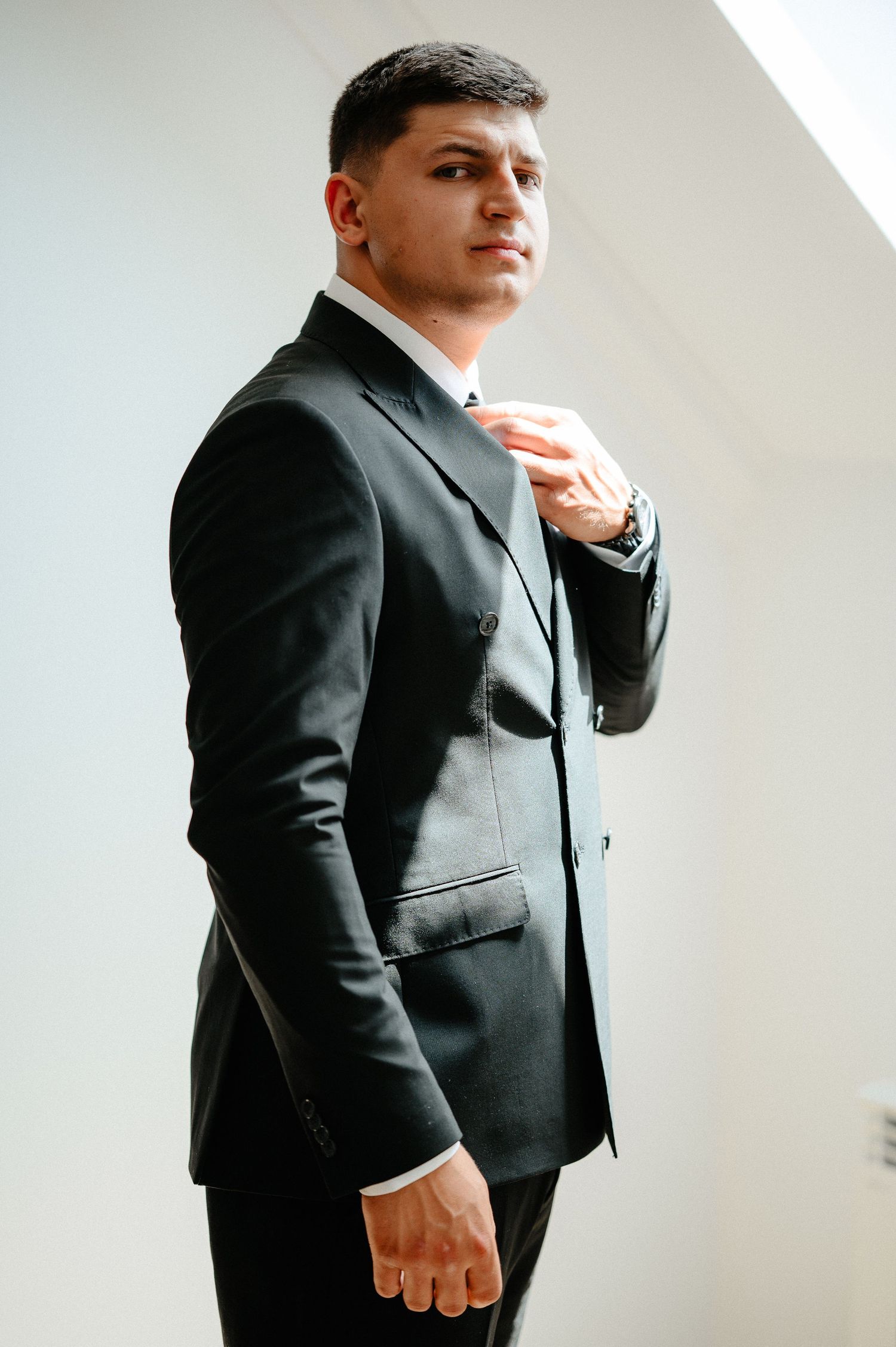 Groom adjusting tie in fitted black suit against white wall.