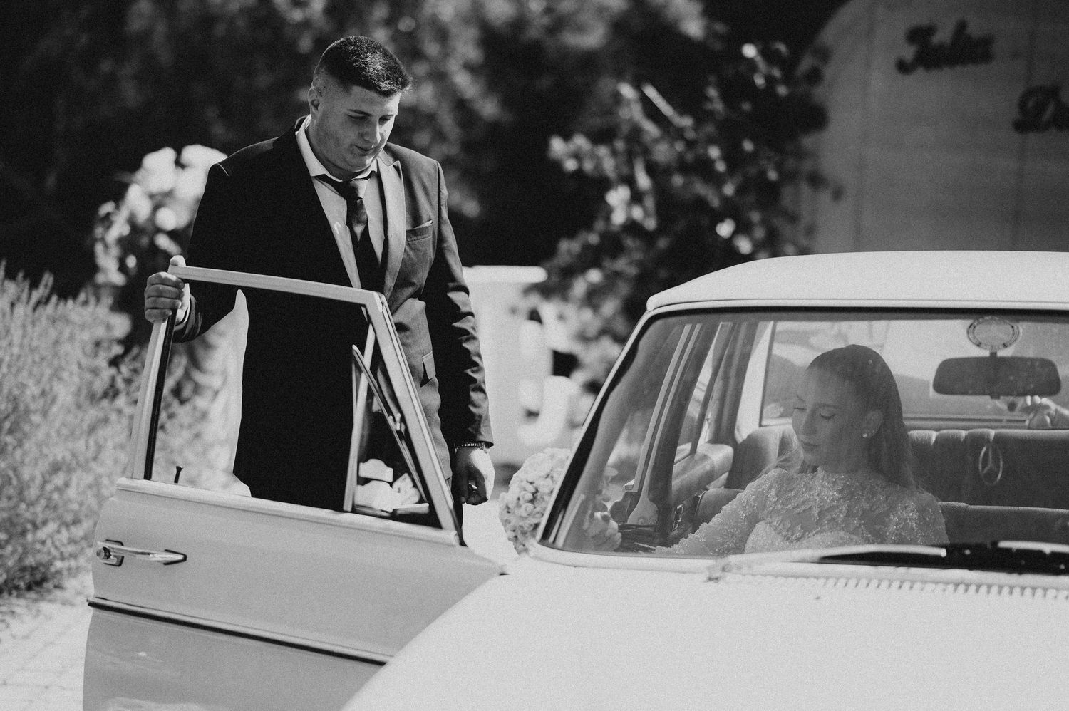 Groom opening car door for bride arriving in vintage white classic car, black and white photo.