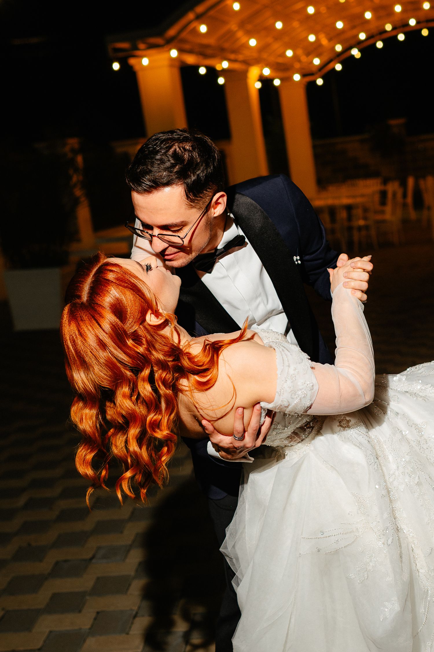 A groom in a tuxedo dips his red-haired bride in a white gown during their romantic first dance under string lights.